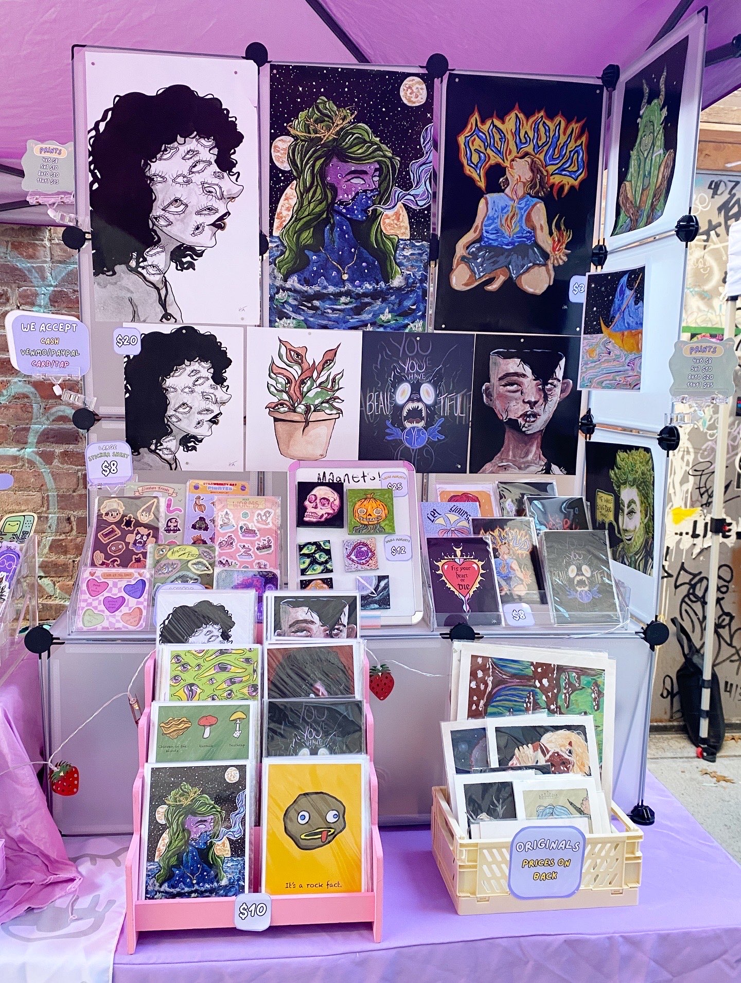 Display of colorful artwork and illustrations on a stand, including portraits, abstract designs, and small cards, with a pink tablecloth.
