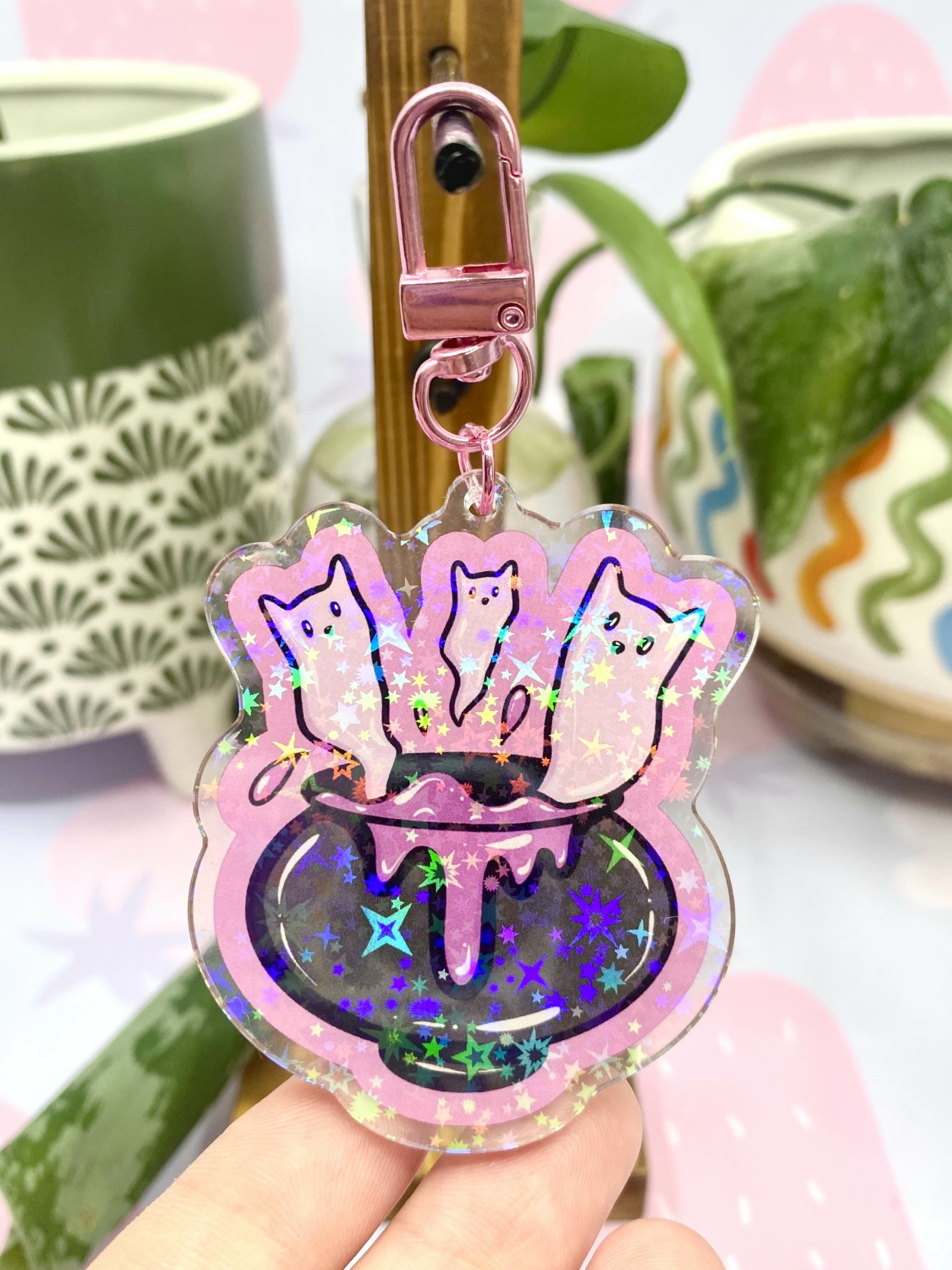 Keychain with three cat figures in a cauldron, glitter and holographic effects, held by a hand, plants in background.