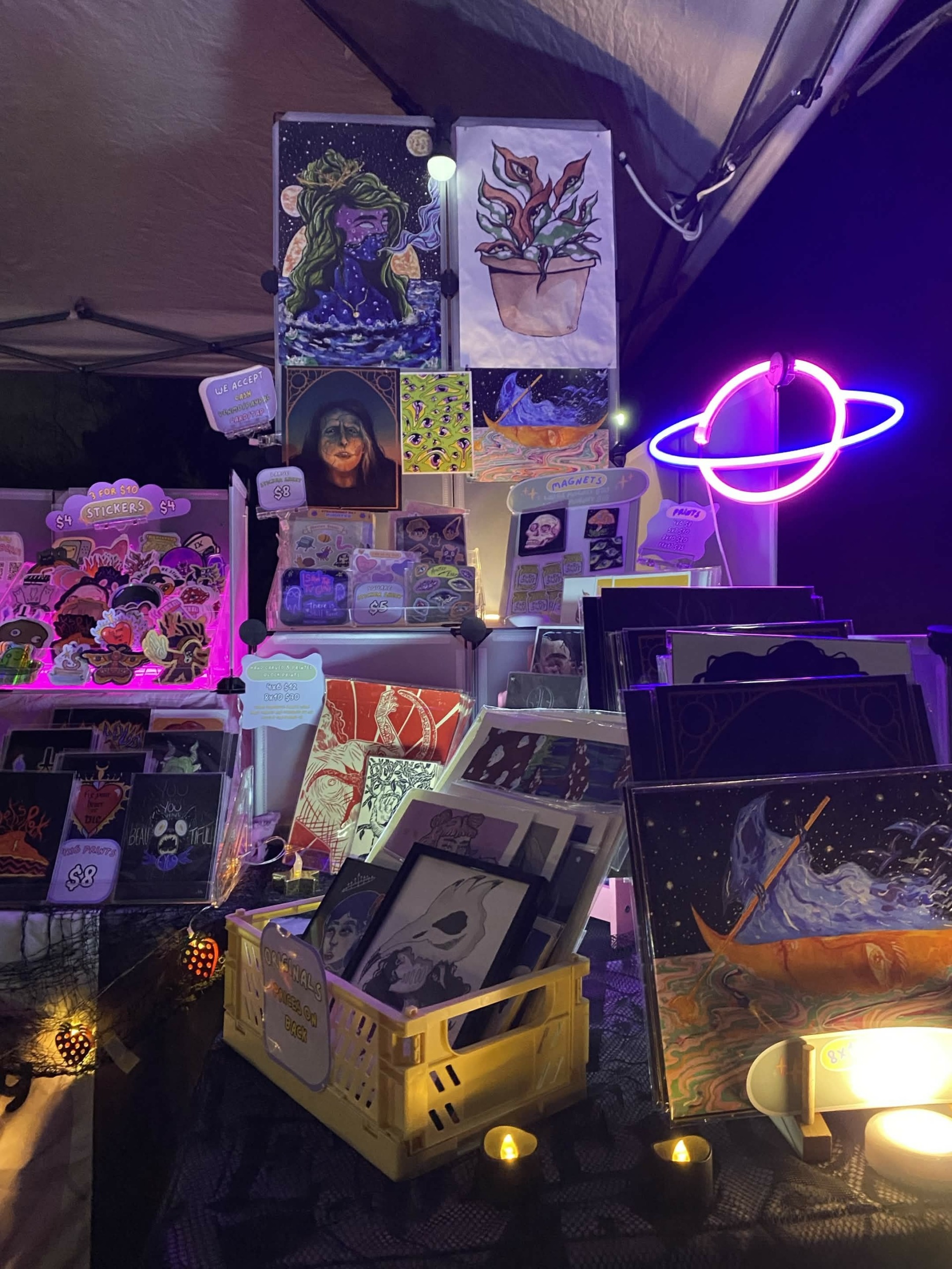 Display of artwork, postcards, and decorative items on a table with a neon planet light in the background.