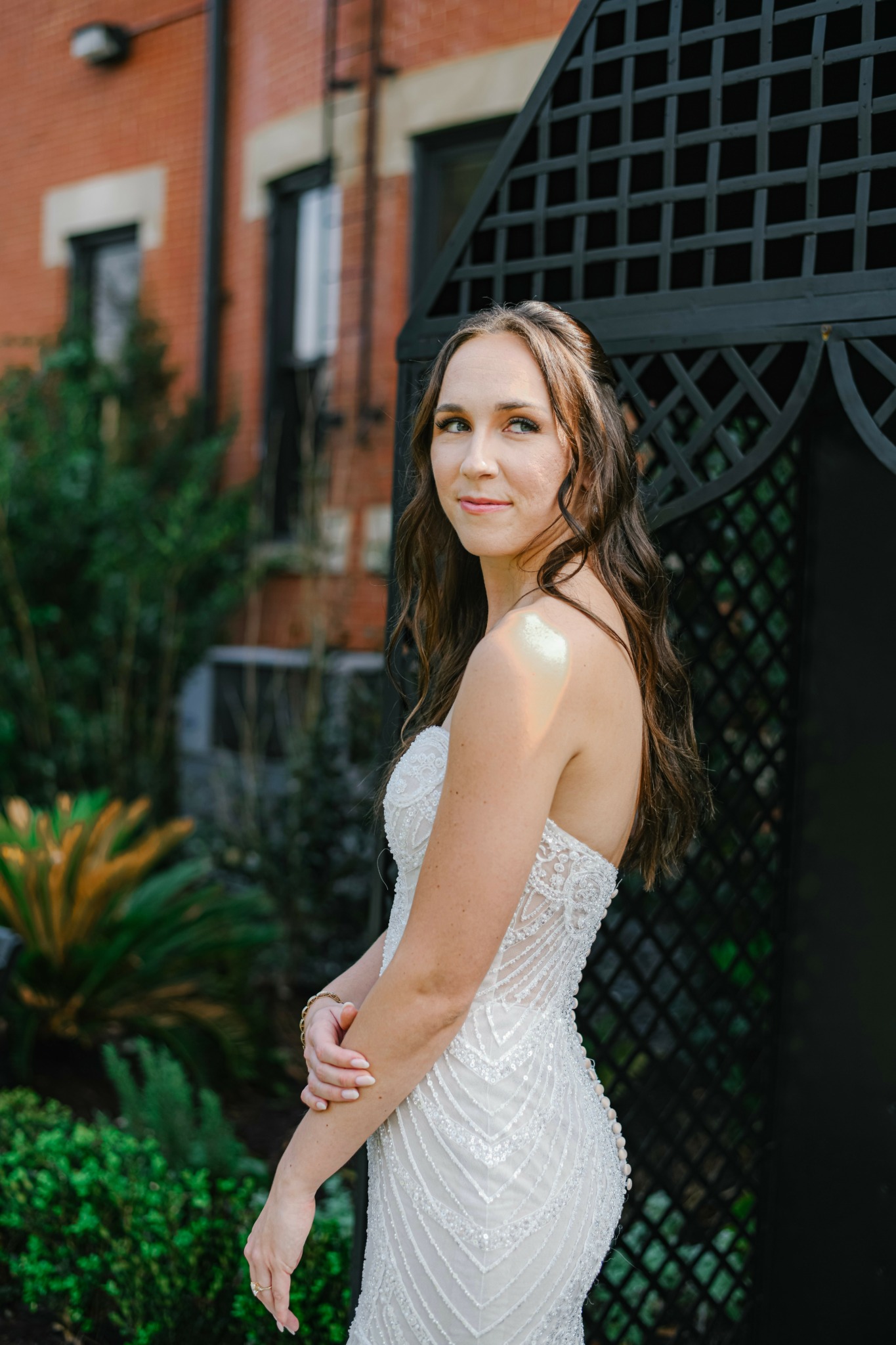 Young woman with long brown hair in a white dress standing outdoors near a black lattice structure.