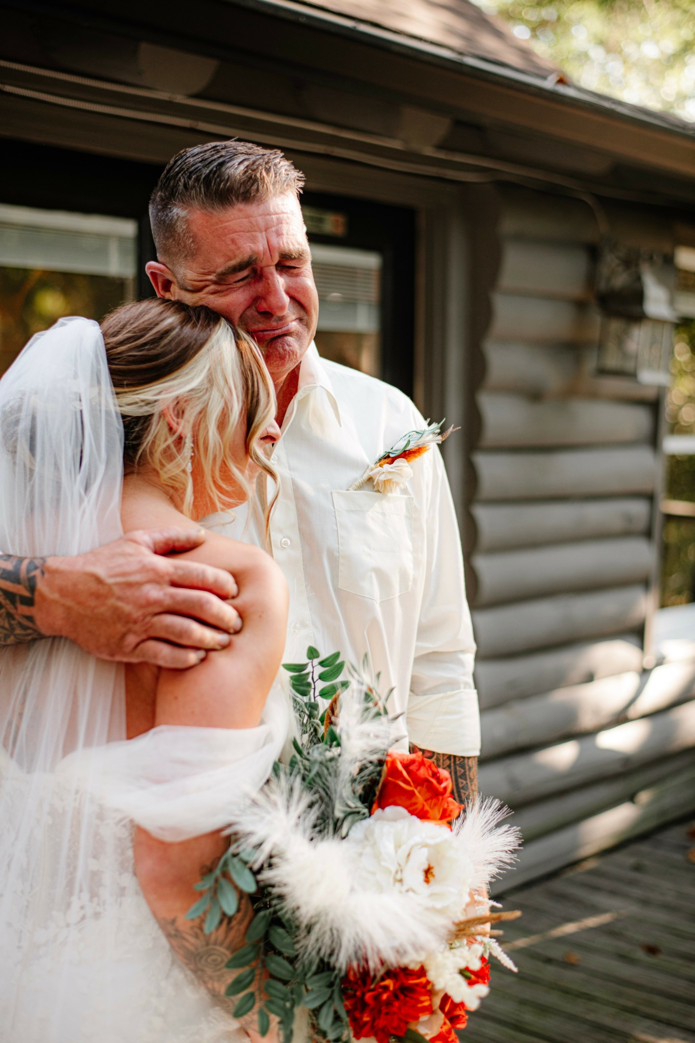 A bride and an older man embrace outdoors, the bride holding a bouquet with red and white flowers and greenery.