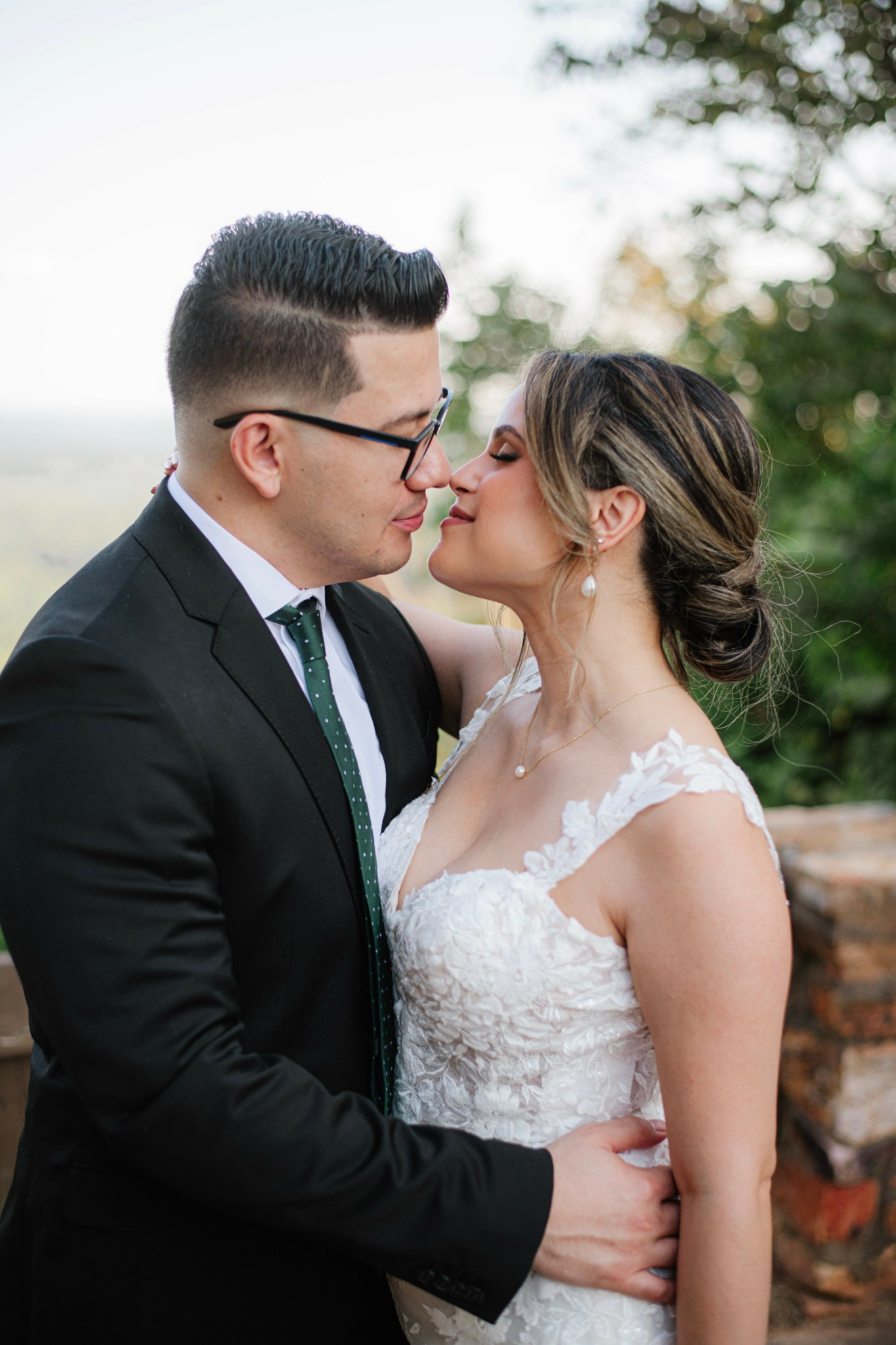 Couple in wedding attire close together outdoors, woman with updo hairstyle, man with glasses, trees in background.