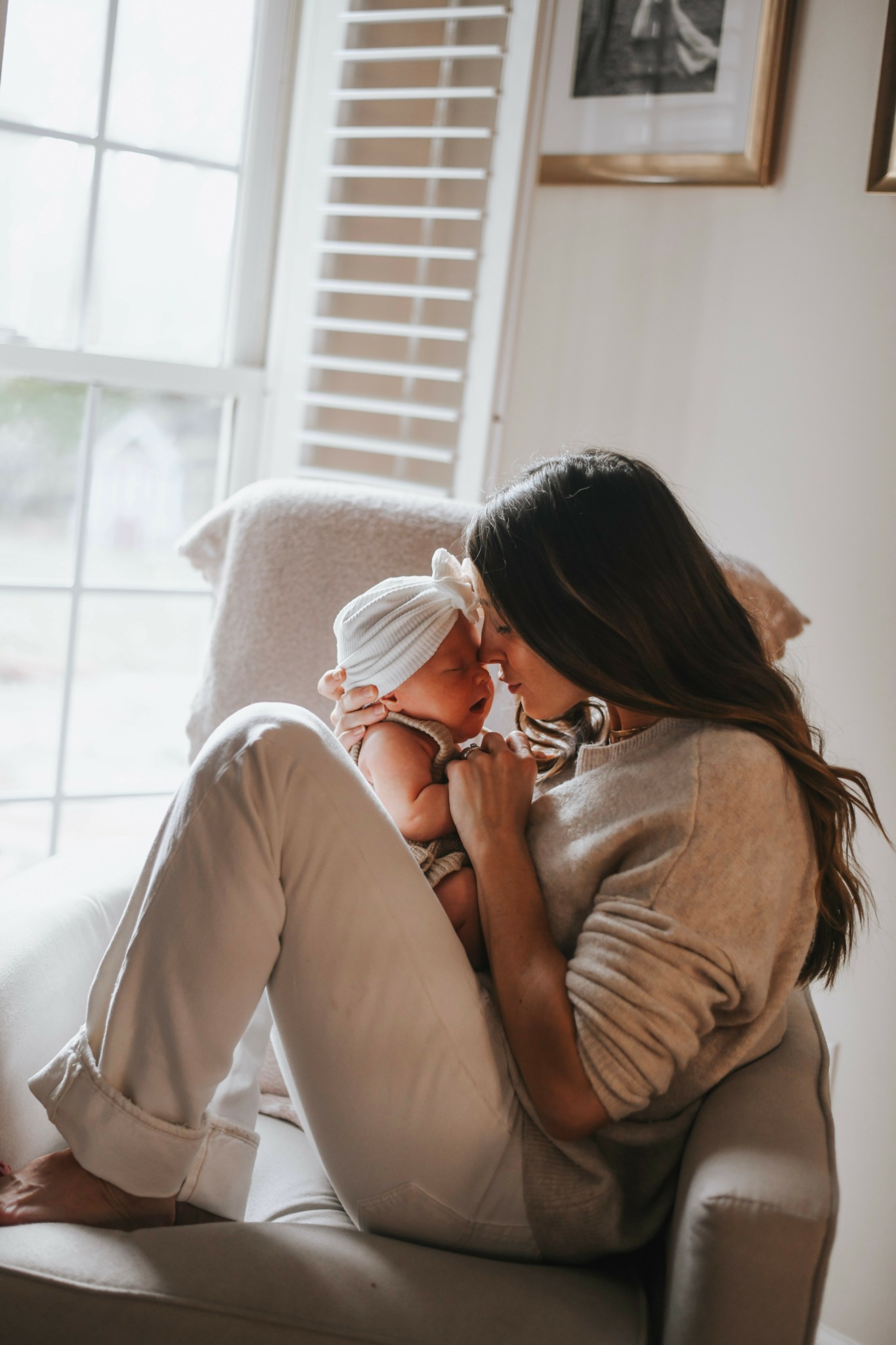 Woman sitting on a chair holding a baby close, both with eyes closed, in a bright room with large window.