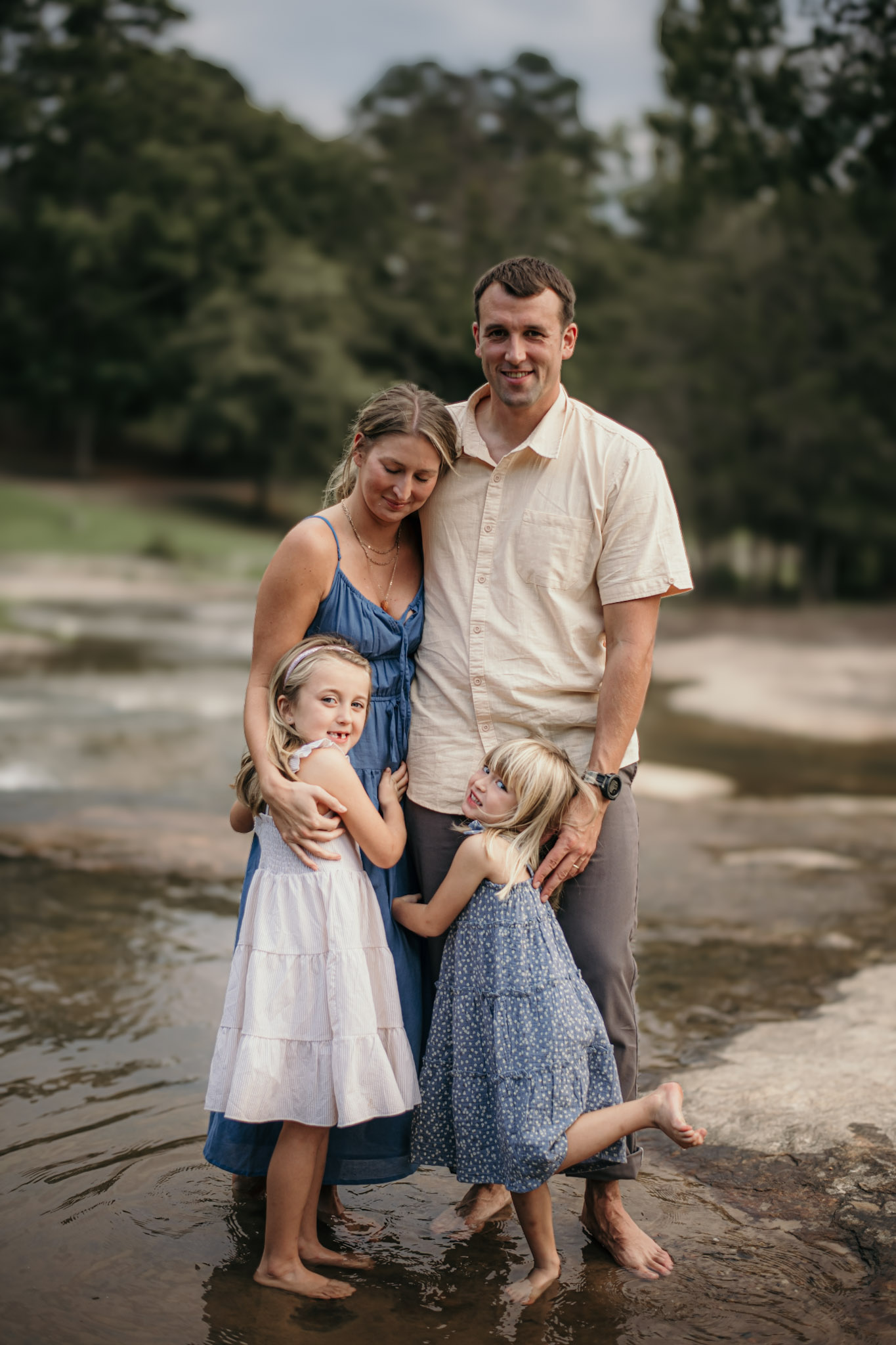 Family of four standing in shallow water outdoors, smiling, with trees in background.