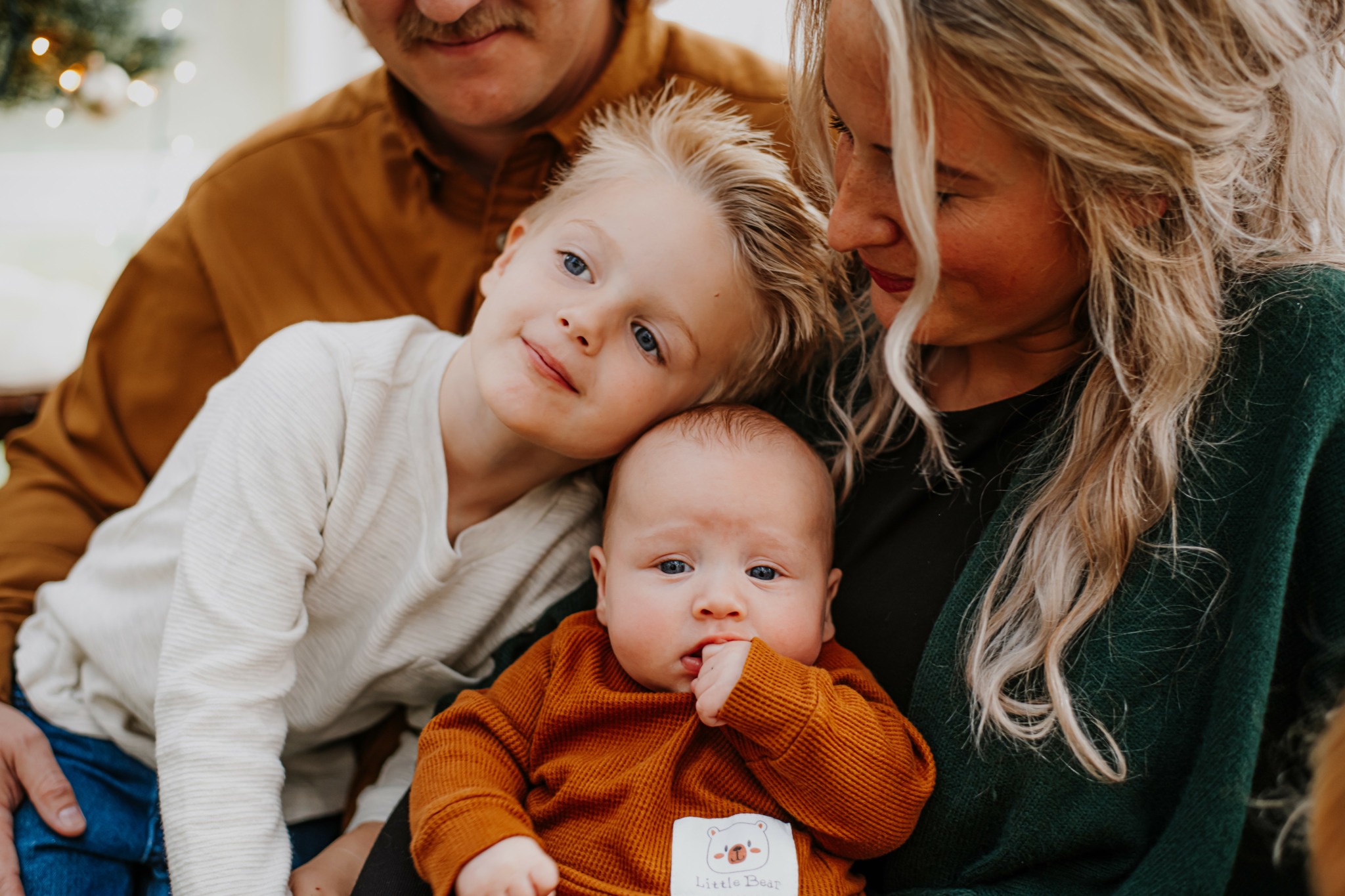 Two children and two adults close together indoors, with the children leaning on the woman with long blonde hair.