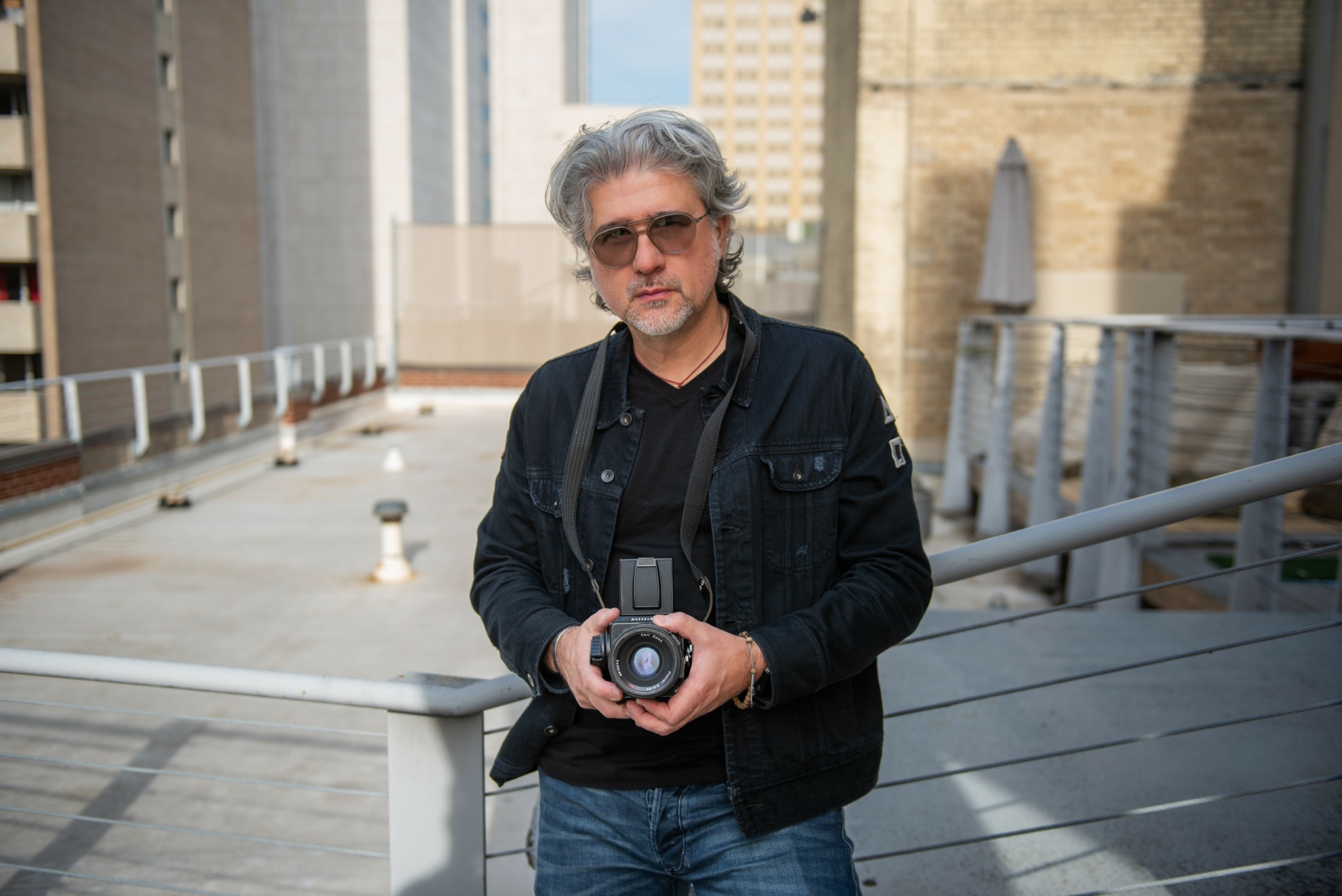 Man with gray hair and sunglasses holding a camera on an outdoor rooftop with buildings in the background.