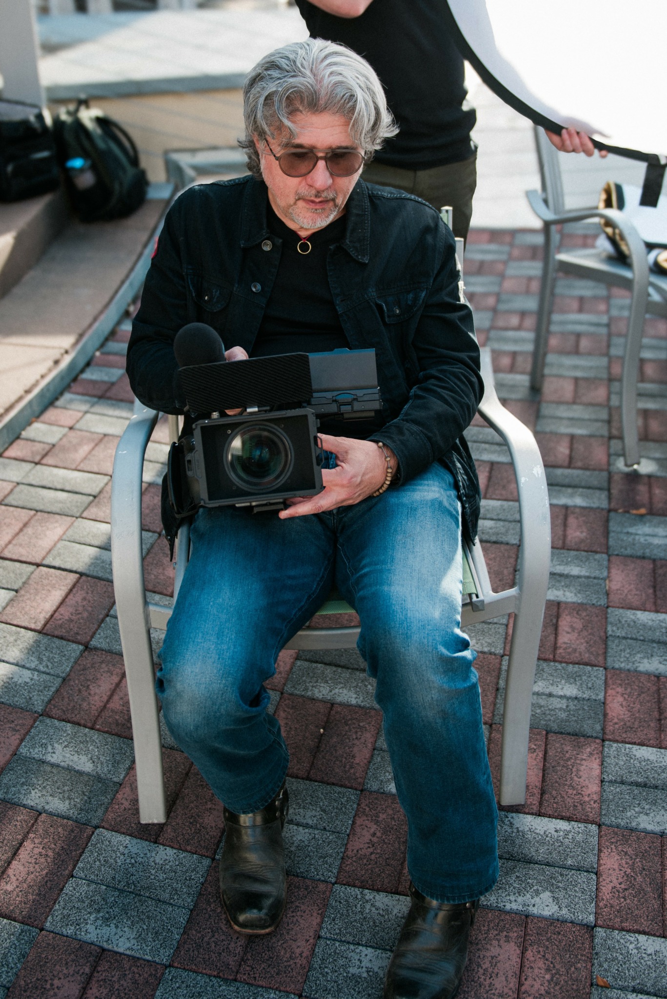 Man with gray hair and sunglasses sitting on a chair holding a camera, outdoors on a brick-paved area.