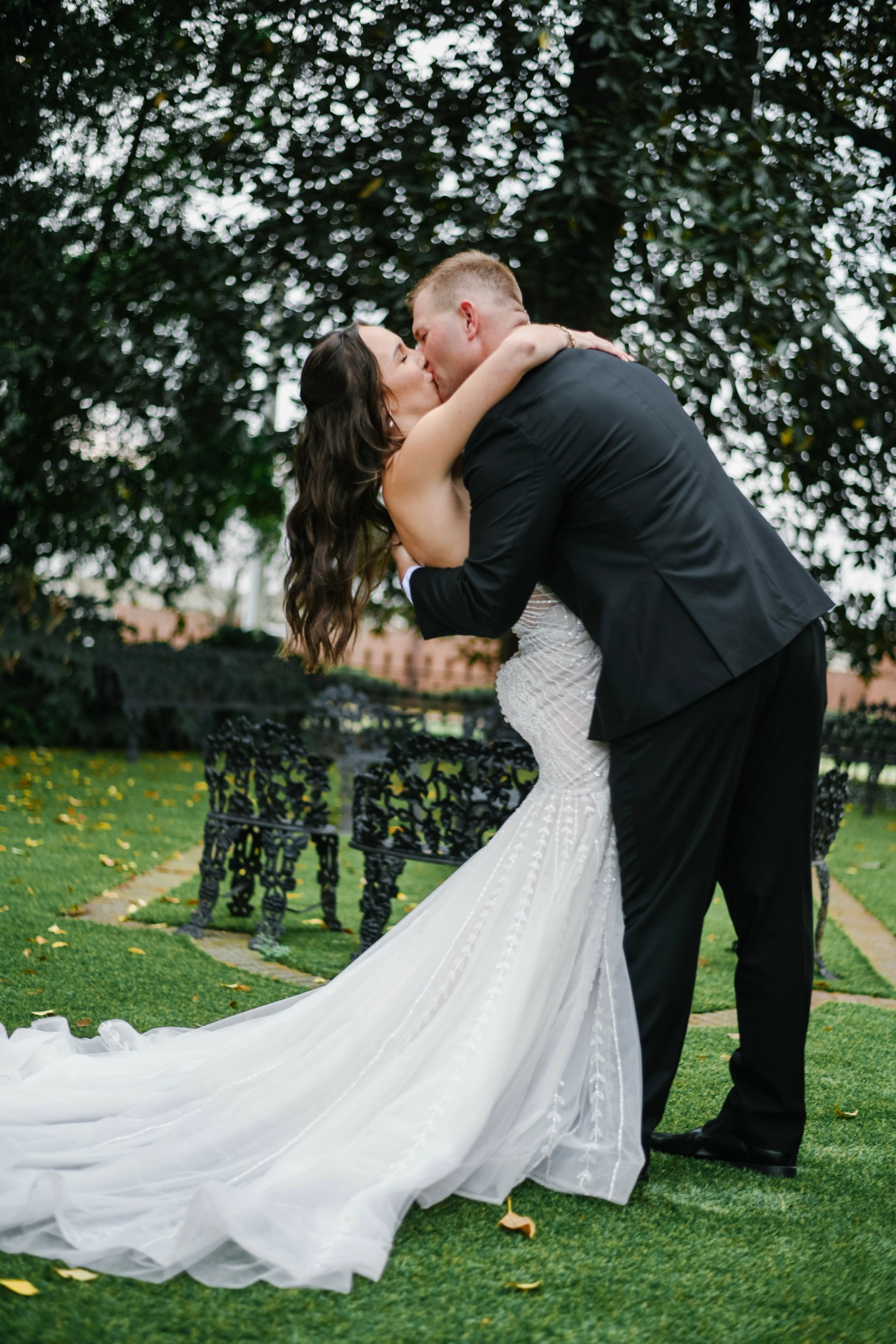 A couple in wedding attire kissing outdoors with trees and a black bench in the background.