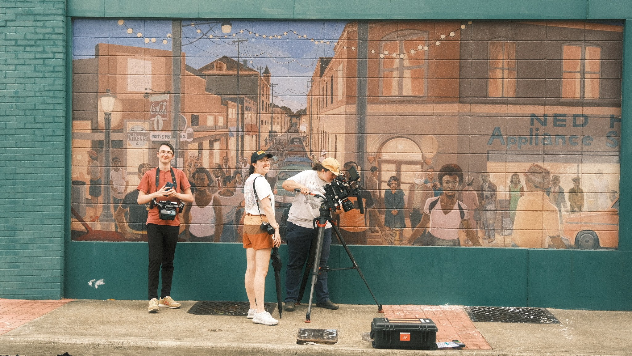 Four people stand outside a large window reflecting buildings and street scene, with cameras and equipment nearby.