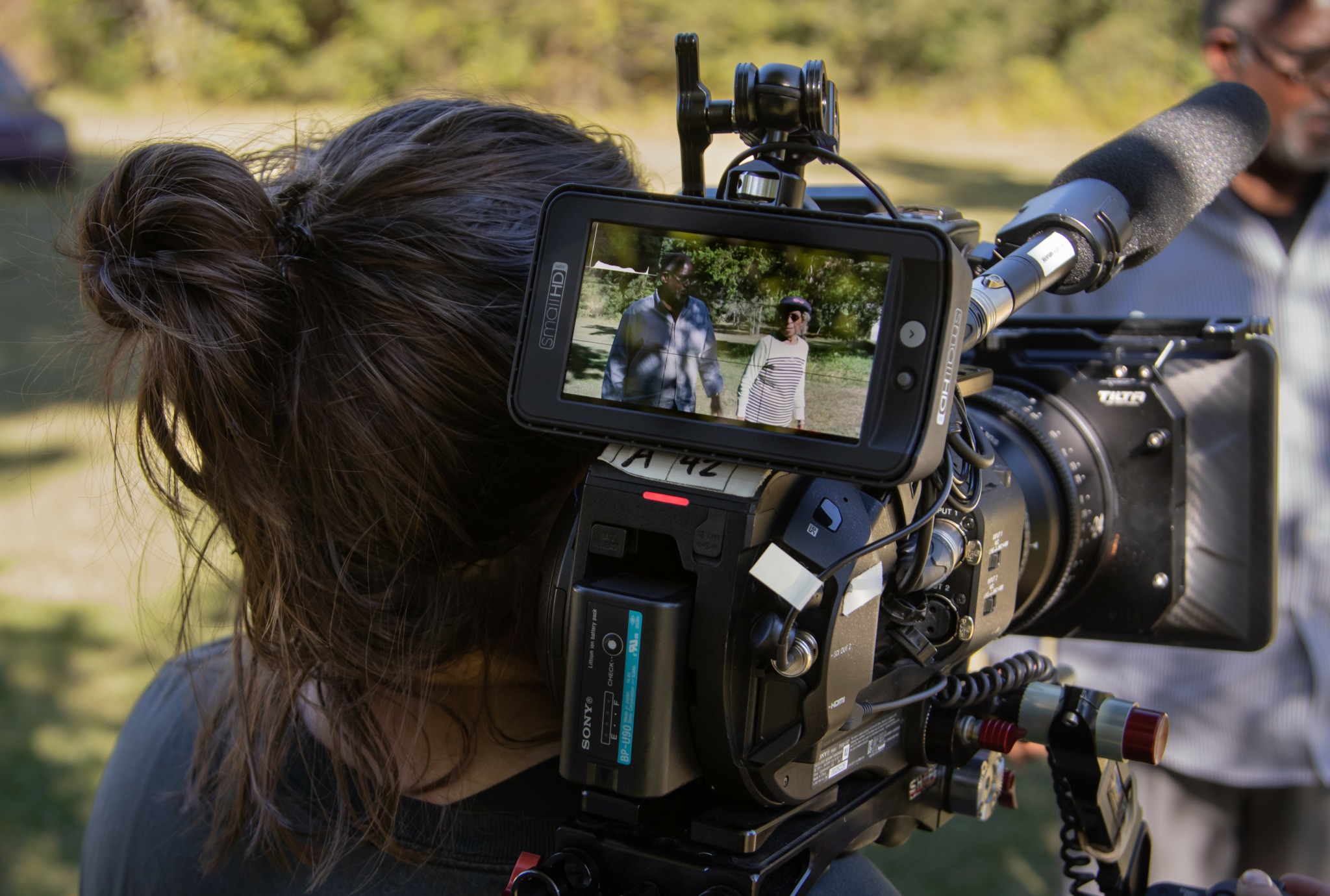 Person with dark hair operating a professional video camera outdoors, with a screen showing two people in the background.