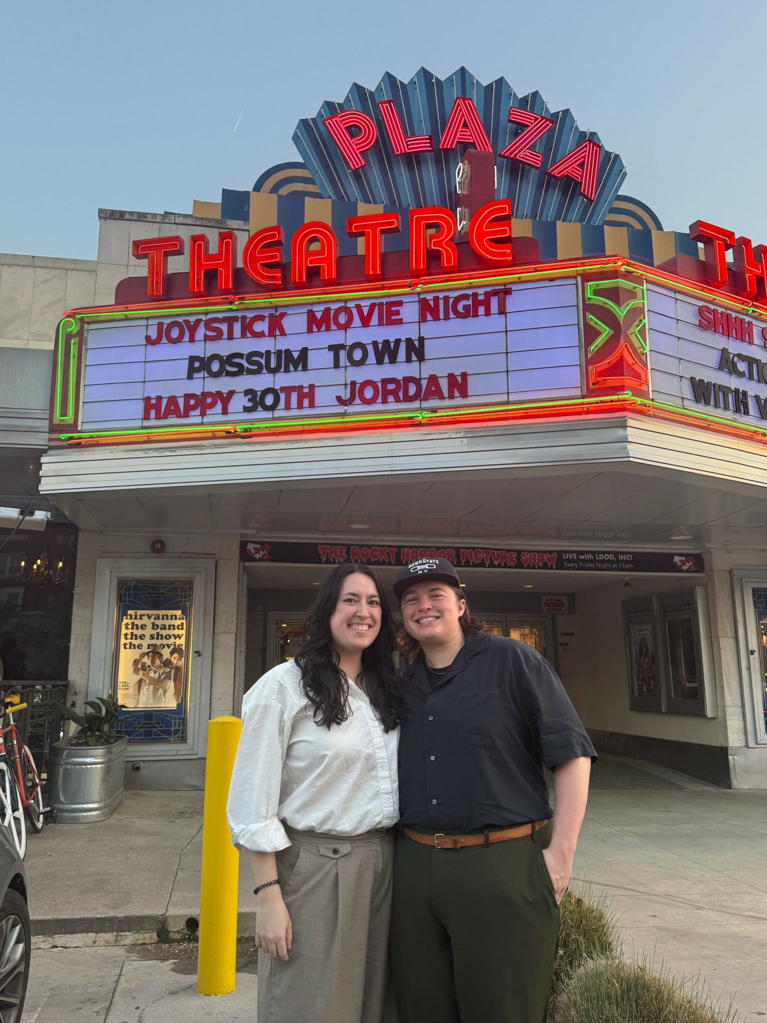 Two women stand outside a movie theater with a marquee displaying movie titles and a message. The theater has a colorful sign.