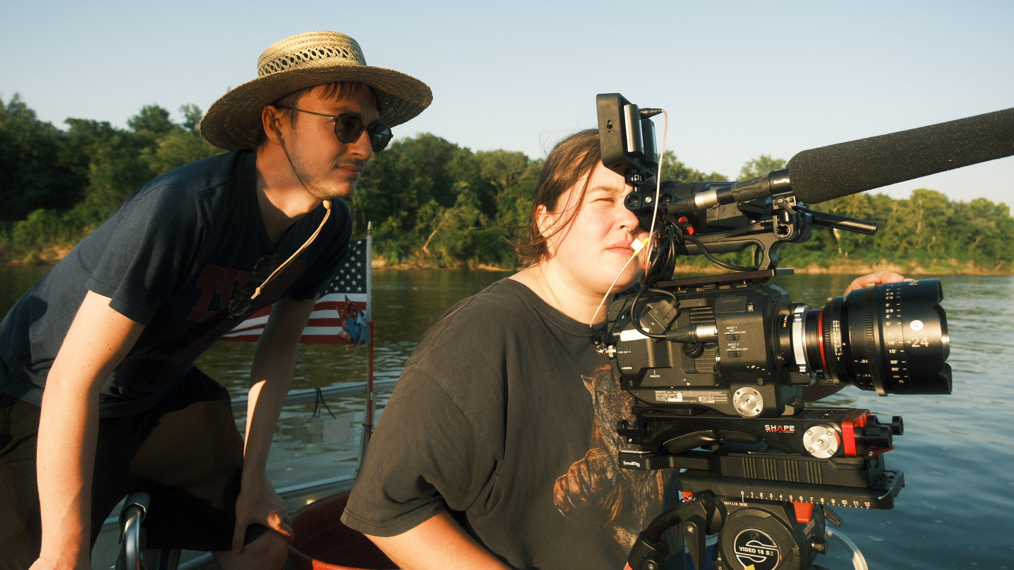 Two women operating a professional camera on a boat near a body of water with trees in the background.
