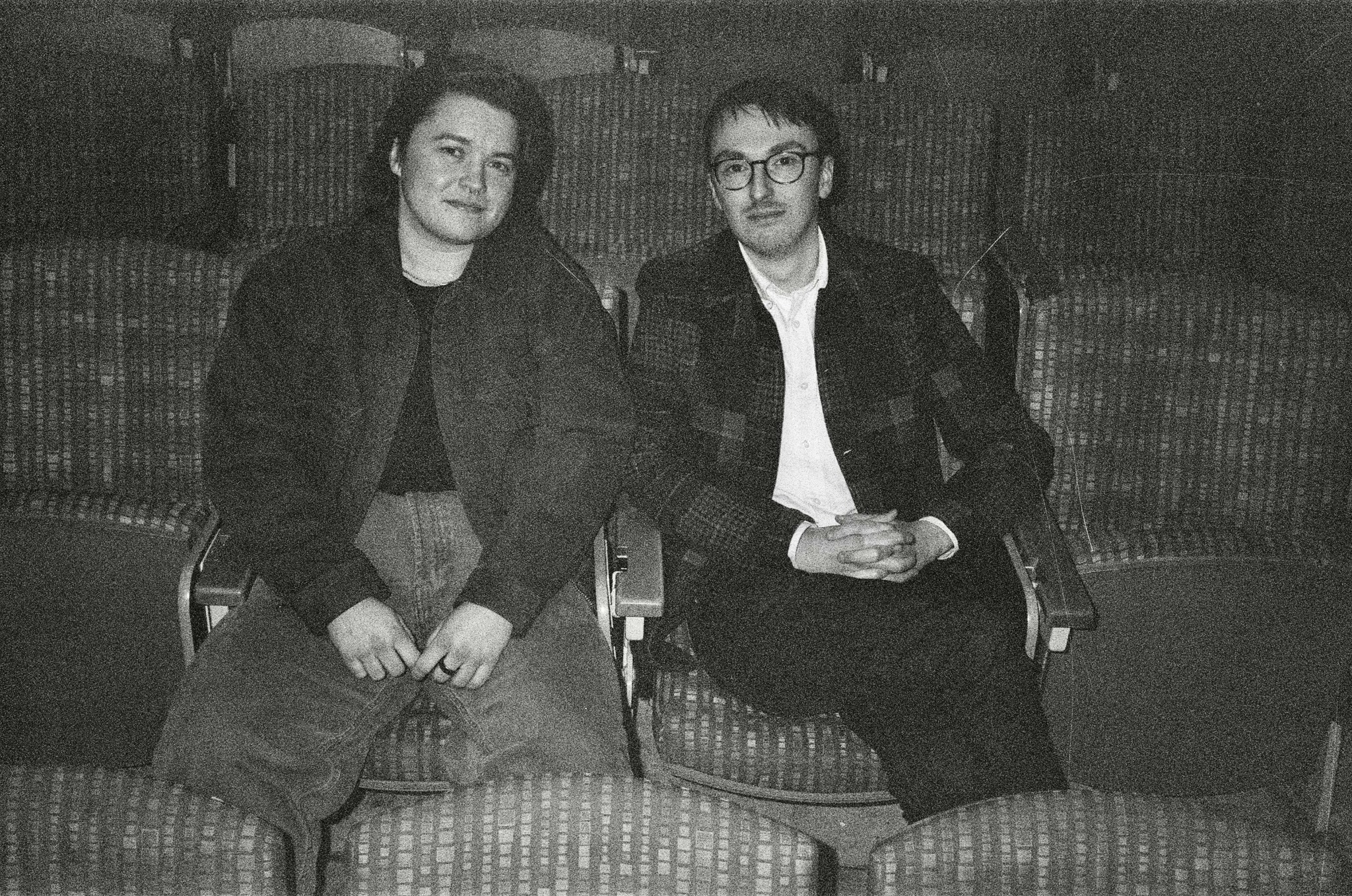 Two people sitting on chairs in a room with patterned walls, facing forward, with empty chairs behind them.