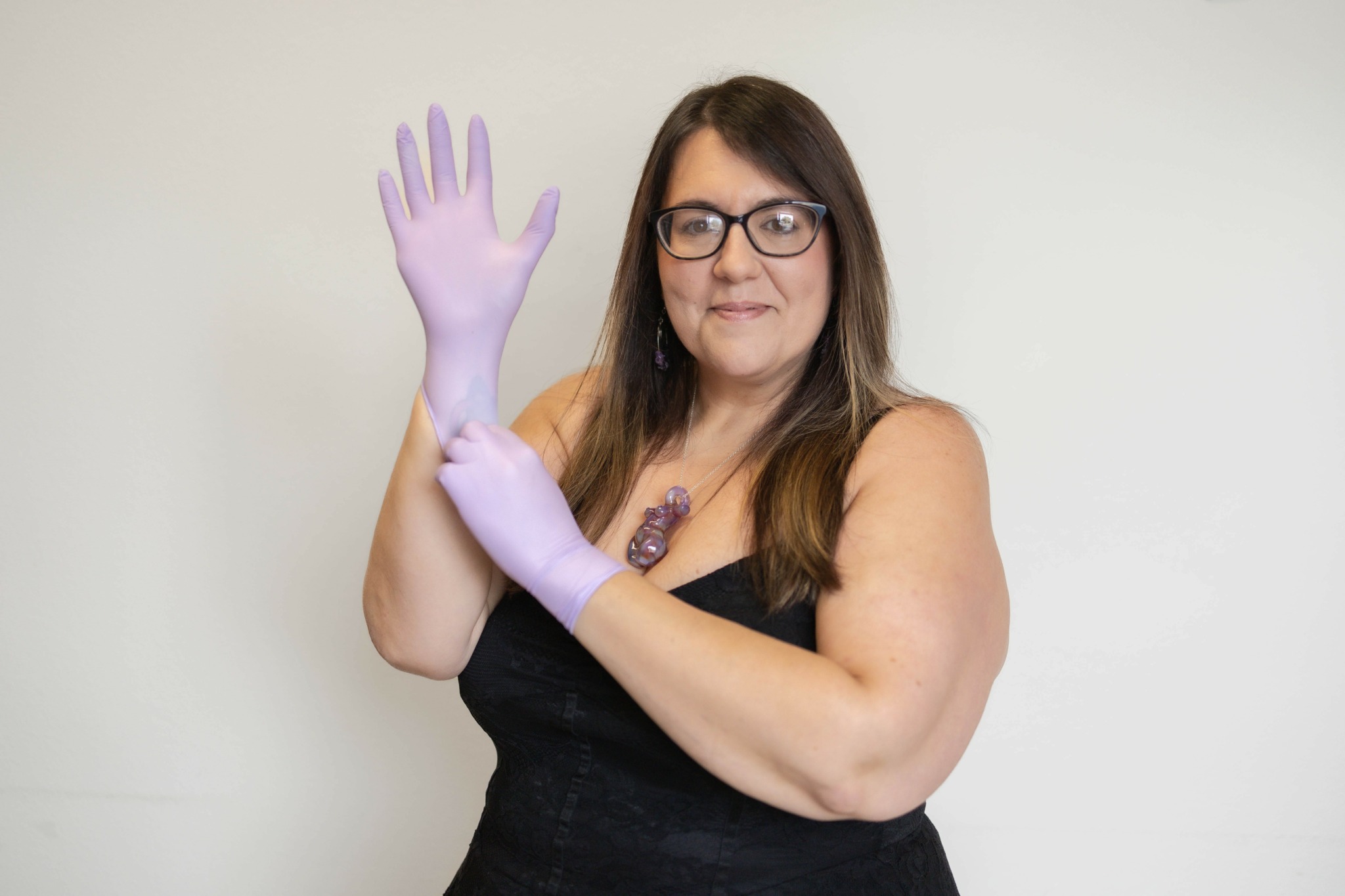 Woman with glasses and long hair wearing a black top, purple gloves, and a necklace, standing against a plain background.