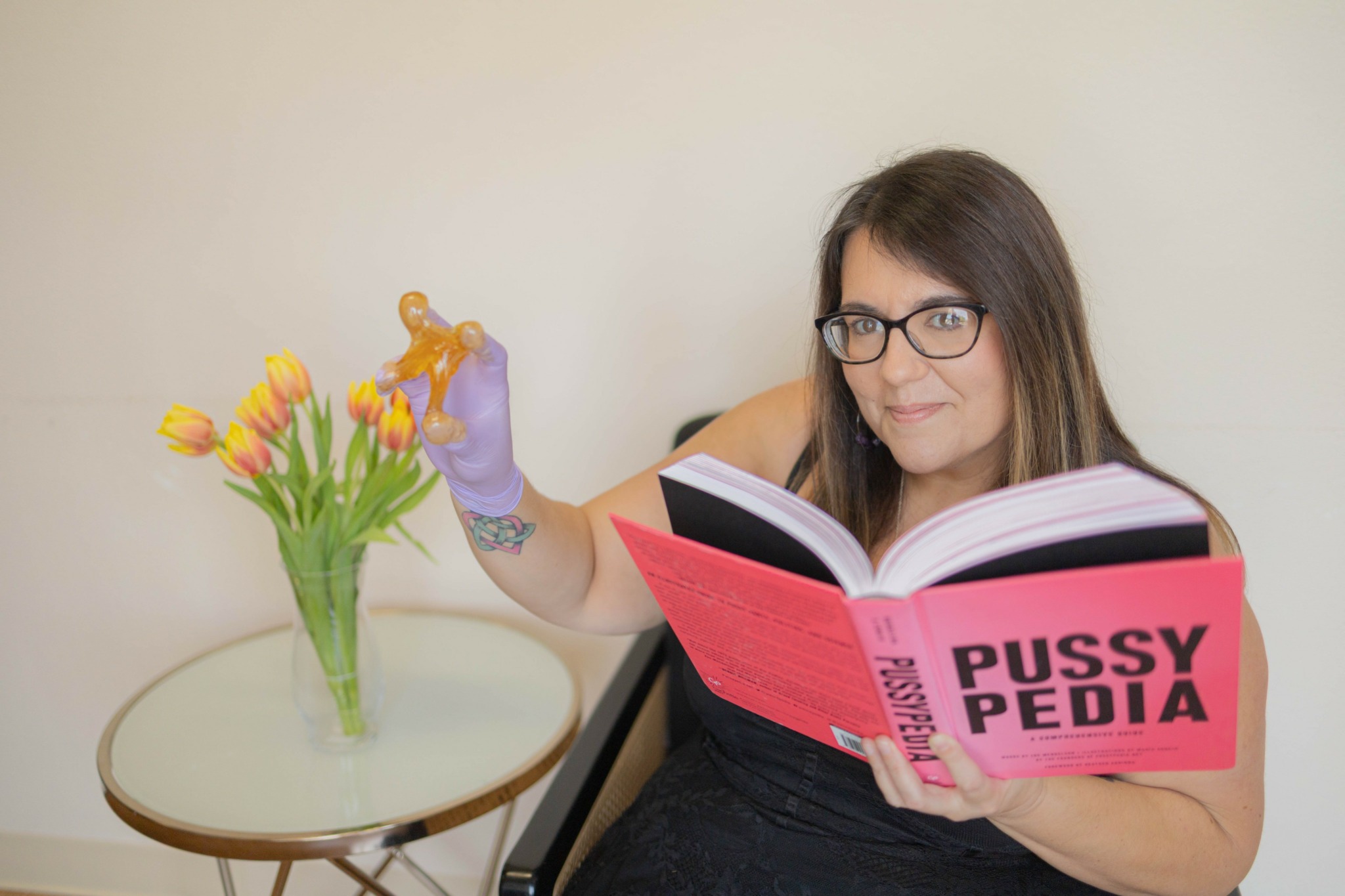 Woman with glasses and tattoos reading a pink book labeled 'Pussy Pedia' while holding a small orange object, sitting at a table with a vase of tulips.