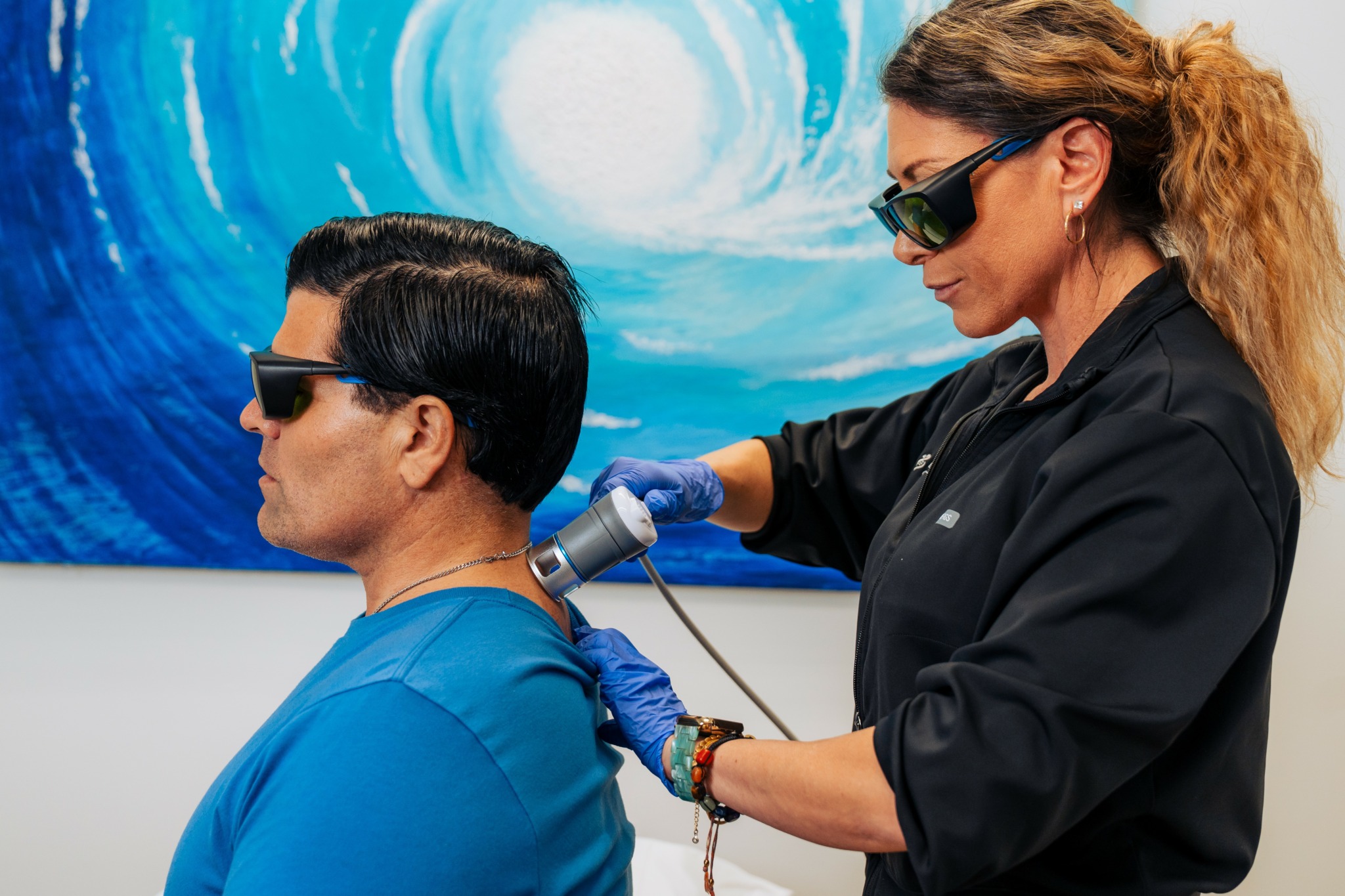 A woman with curly hair and sunglasses performs a cosmetic procedure on a seated woman with short dark hair, wearing a blue top.