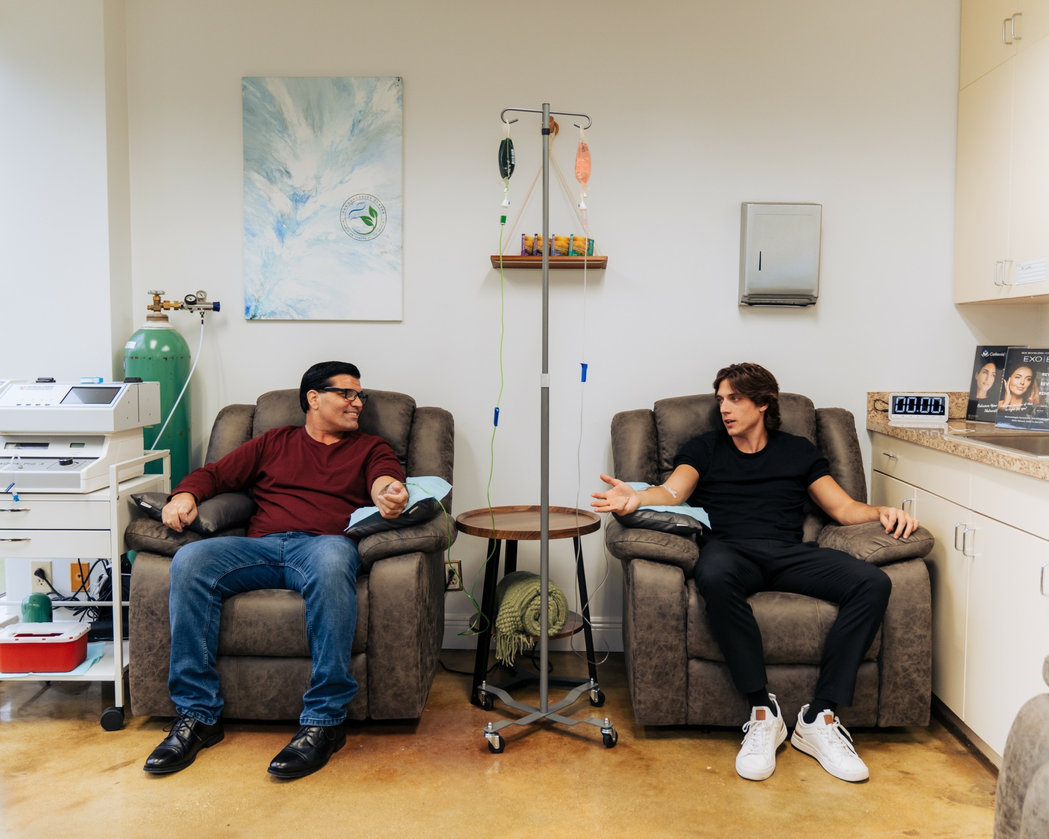 Two people sitting on couches in a medical room, talking, with medical equipment and artwork on the wall.