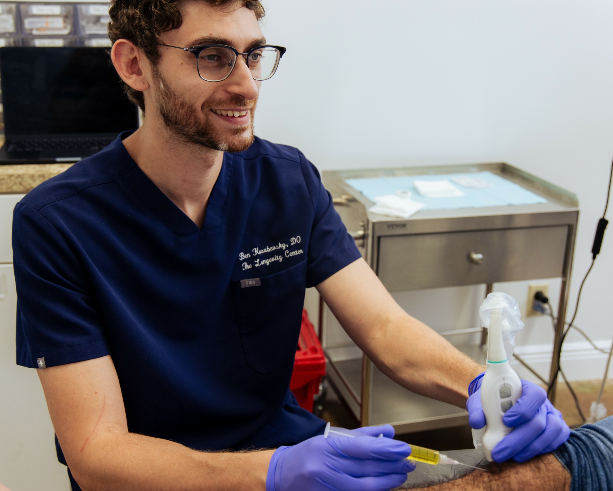 Young man with glasses and beard wearing navy scrubs and gloves, smiling while working with a pipette in a clinical setting.