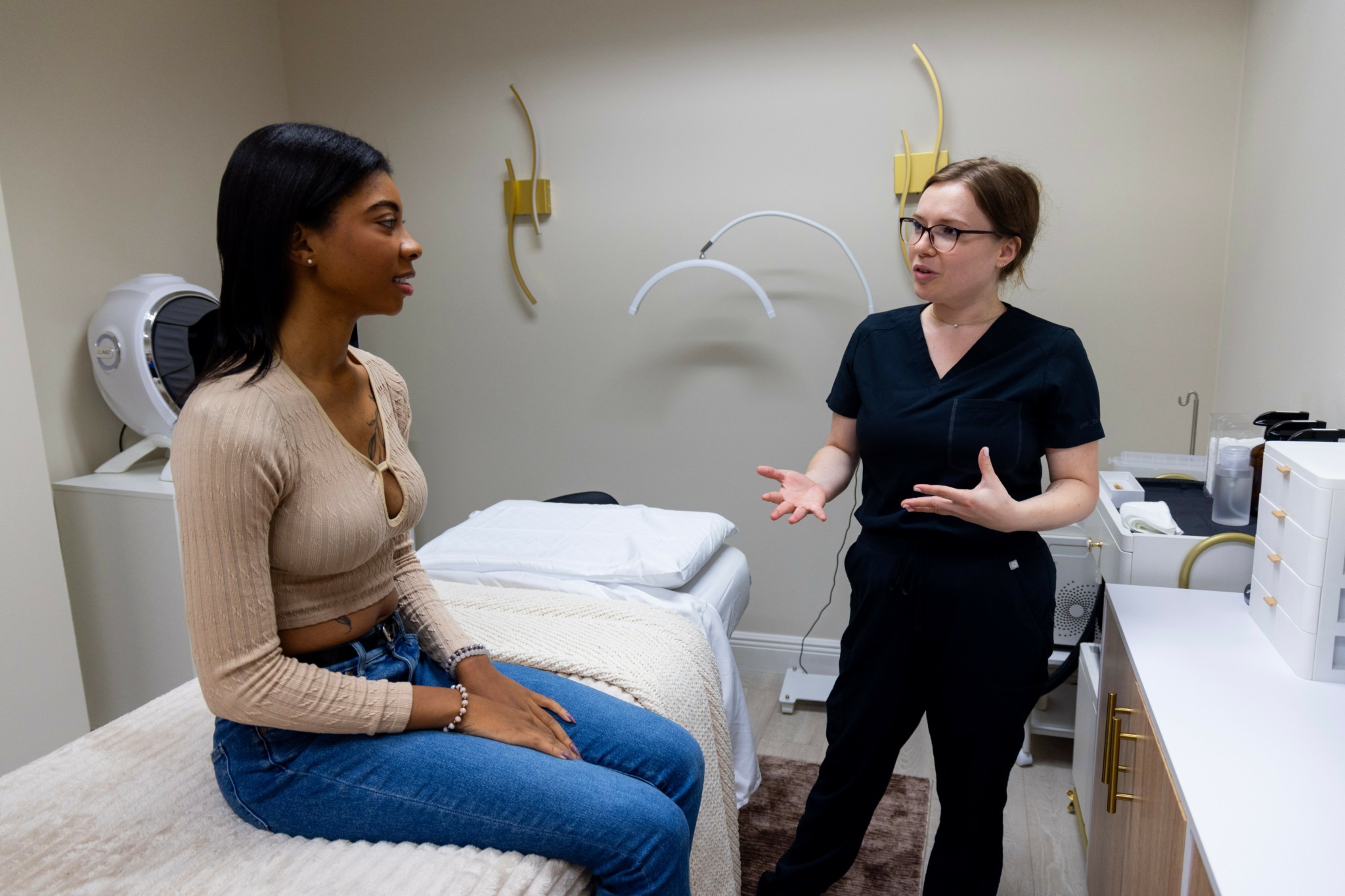 Two women in conversation in a medical examination room with a bed and medical equipment.