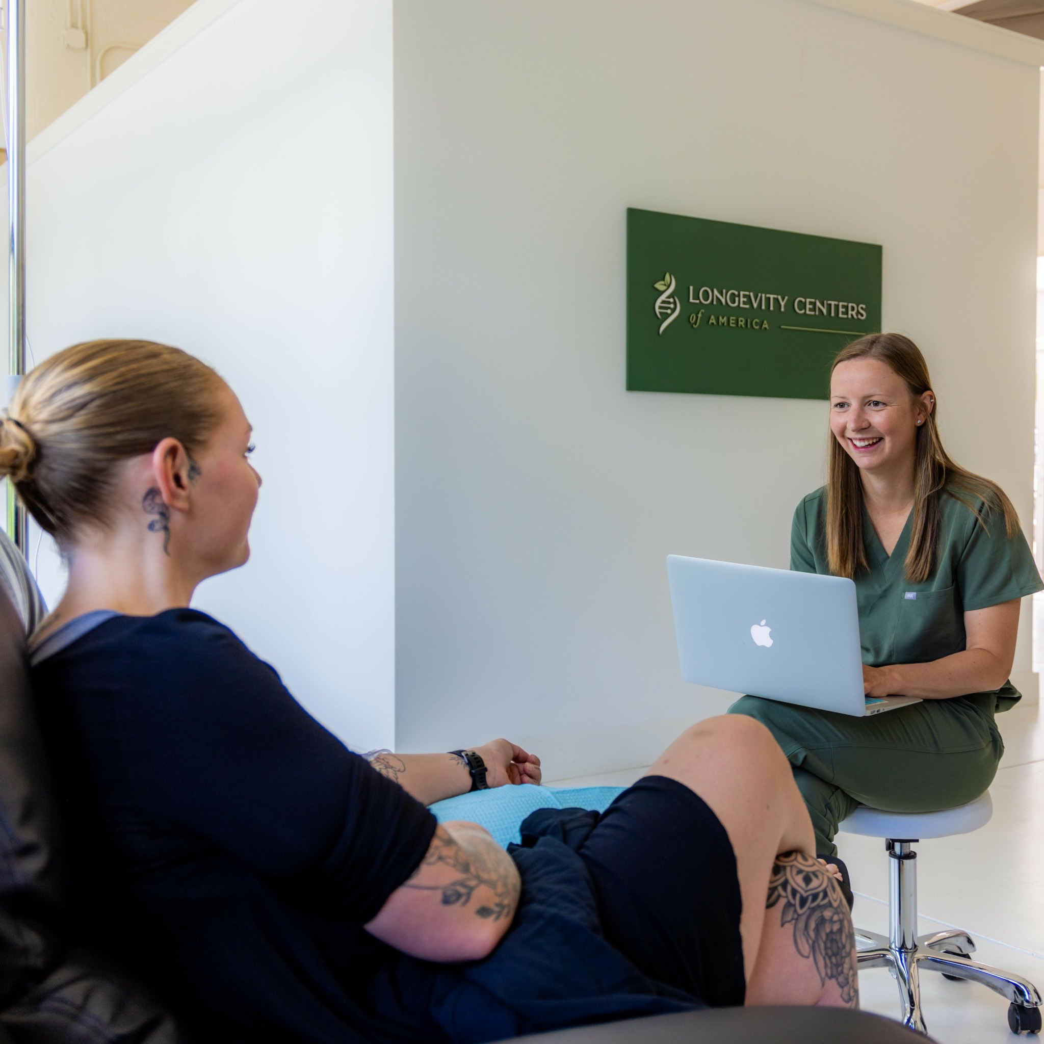 Two women sitting and talking in a medical office with a green sign on the wall.