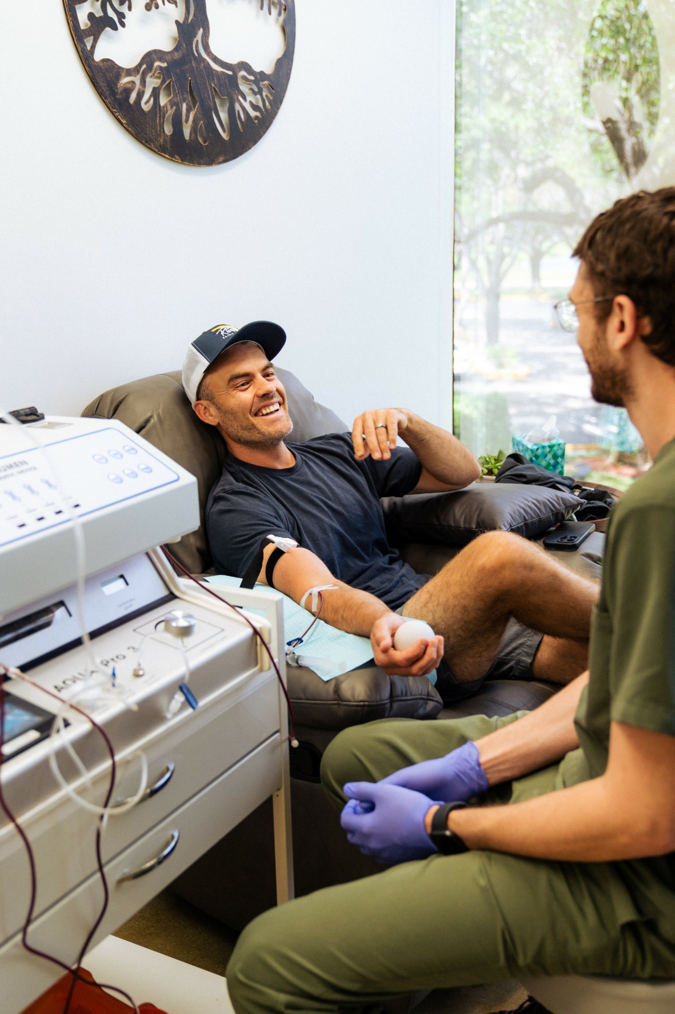 Young man smiling and talking to healthcare worker in a medical office, sitting in a chair with medical equipment nearby.