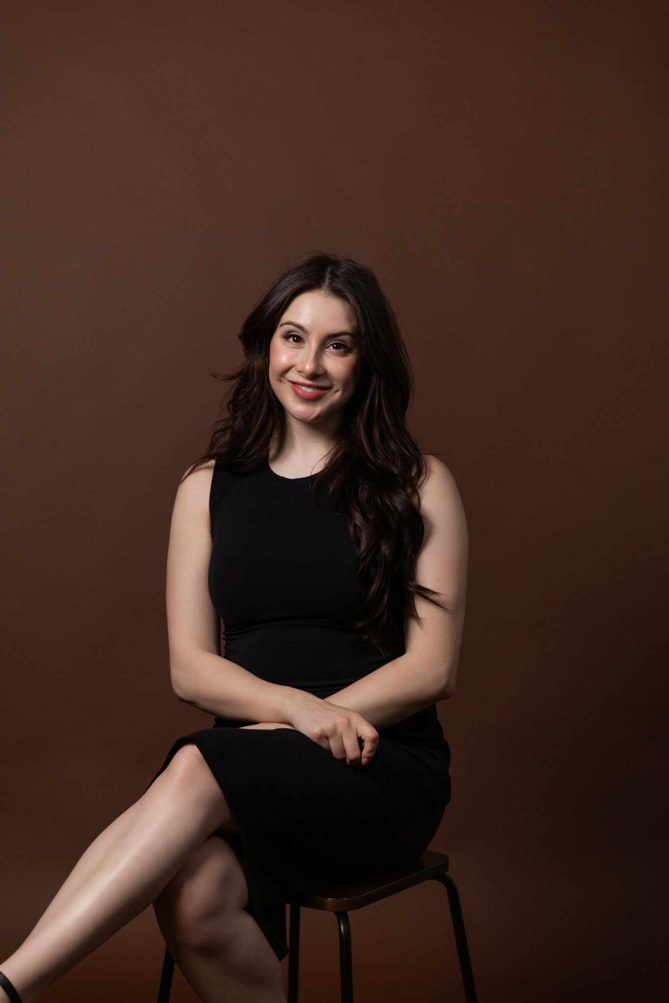 Young woman with long dark hair sitting on a stool against a brown background, smiling, wearing a black sleeveless dress.