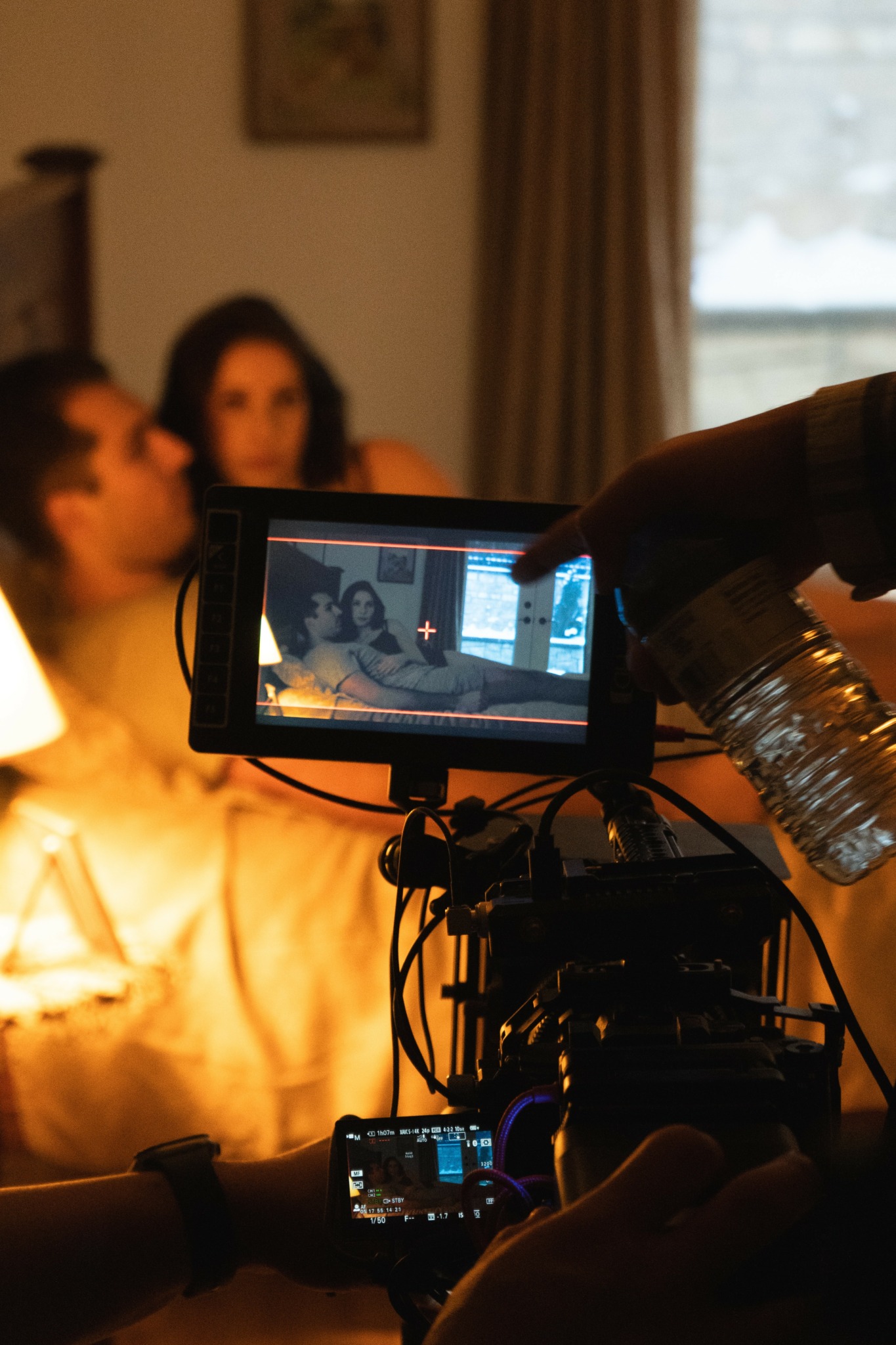 Filmmaker records a scene with two actors sitting on a bed in a dimly lit room, window with curtains in background.