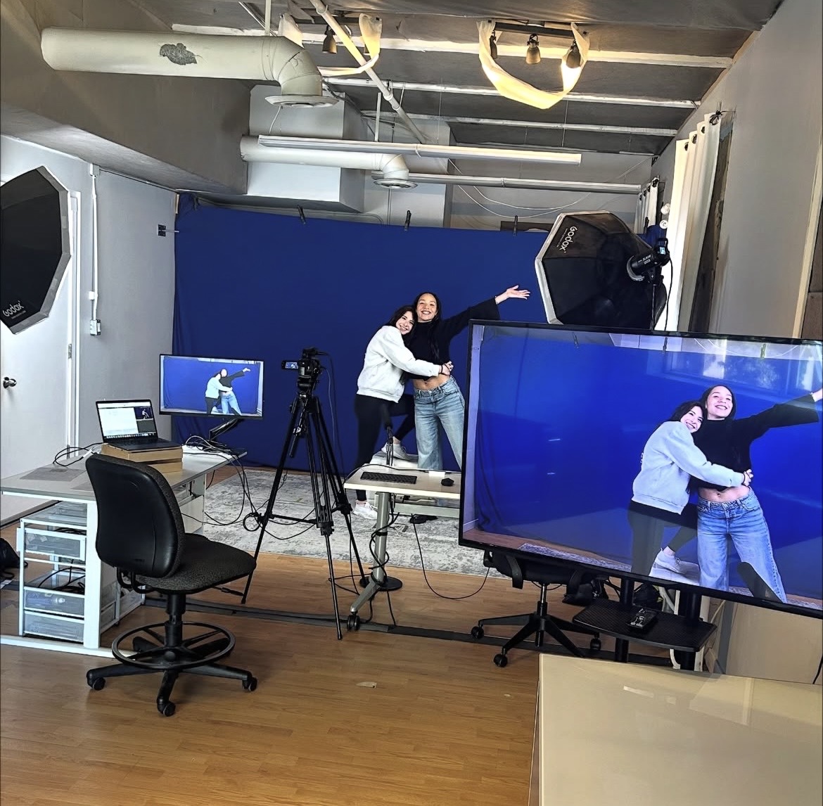 Two people in a studio, one with arm around the other, with camera equipment and monitors showing the scene.