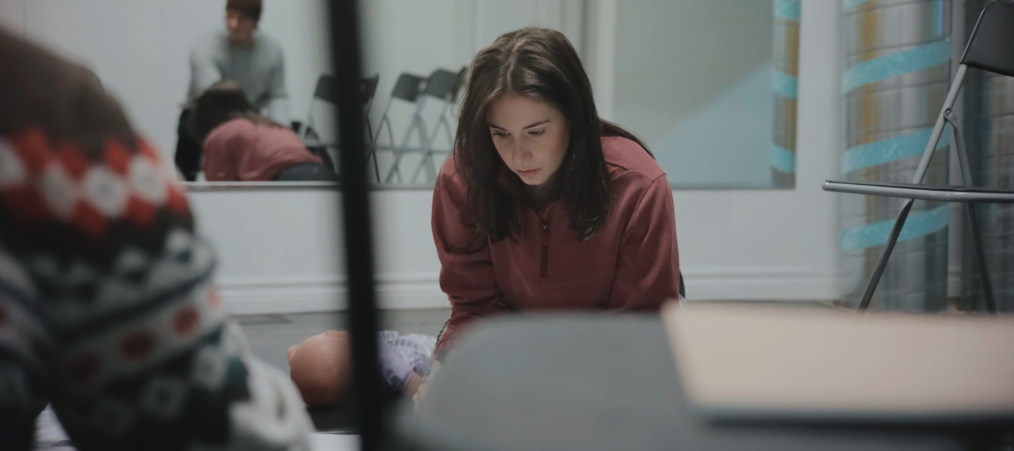 Young woman with long hair looking down, sitting indoors near a window with curtains, in a room with chairs and a mirror.