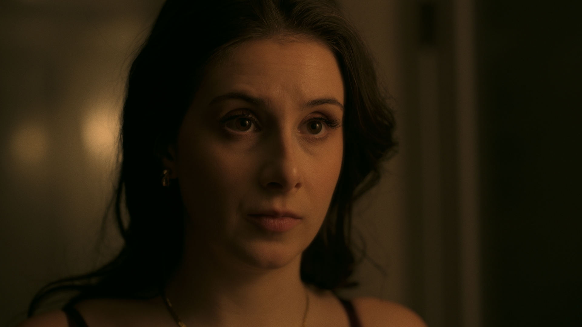 Close-up of a woman with dark hair and a serious expression, indoors with warm lighting.