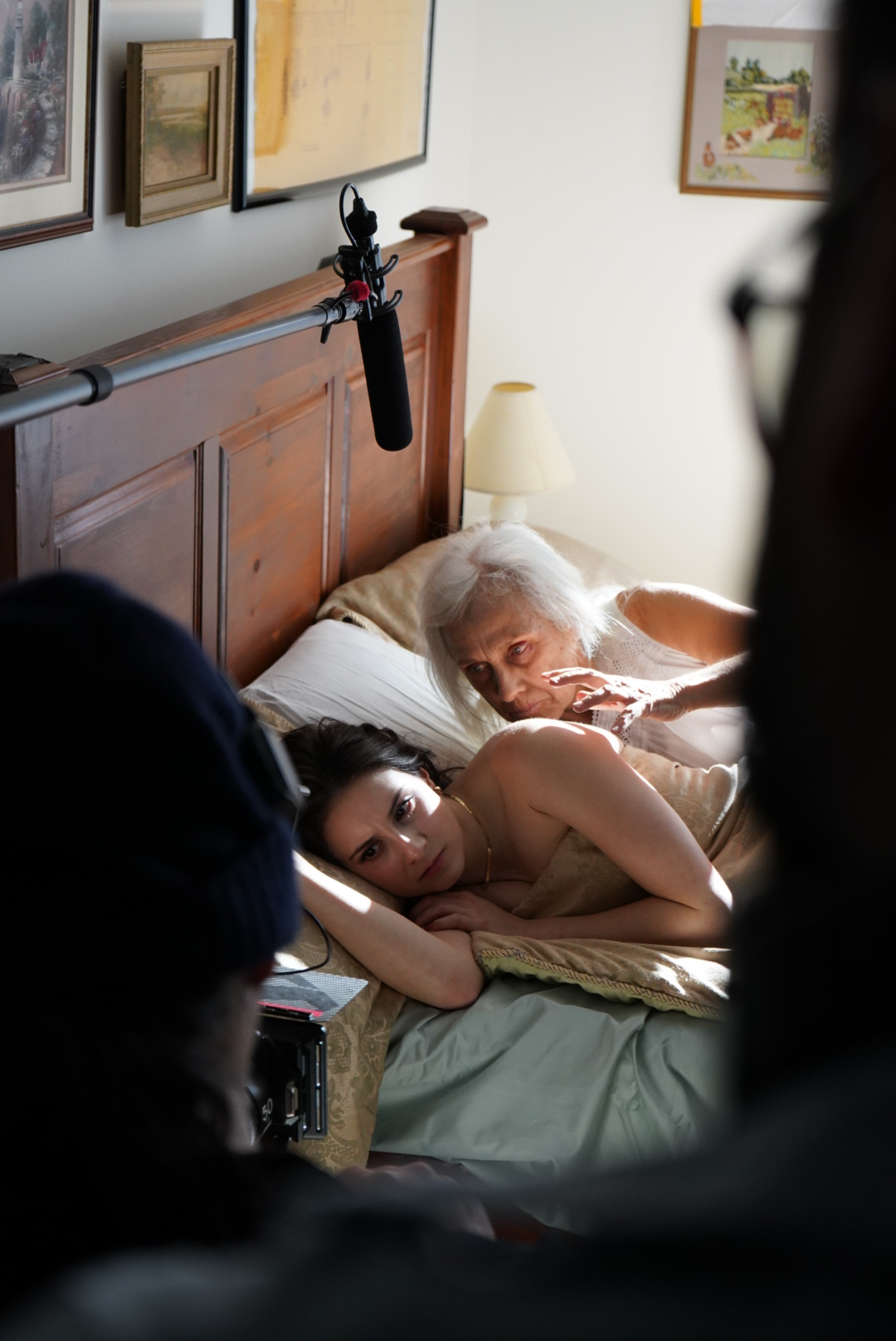 Elderly woman lying in bed with young girl, both wearing face masks, in a bedroom with wooden furniture and framed pictures.