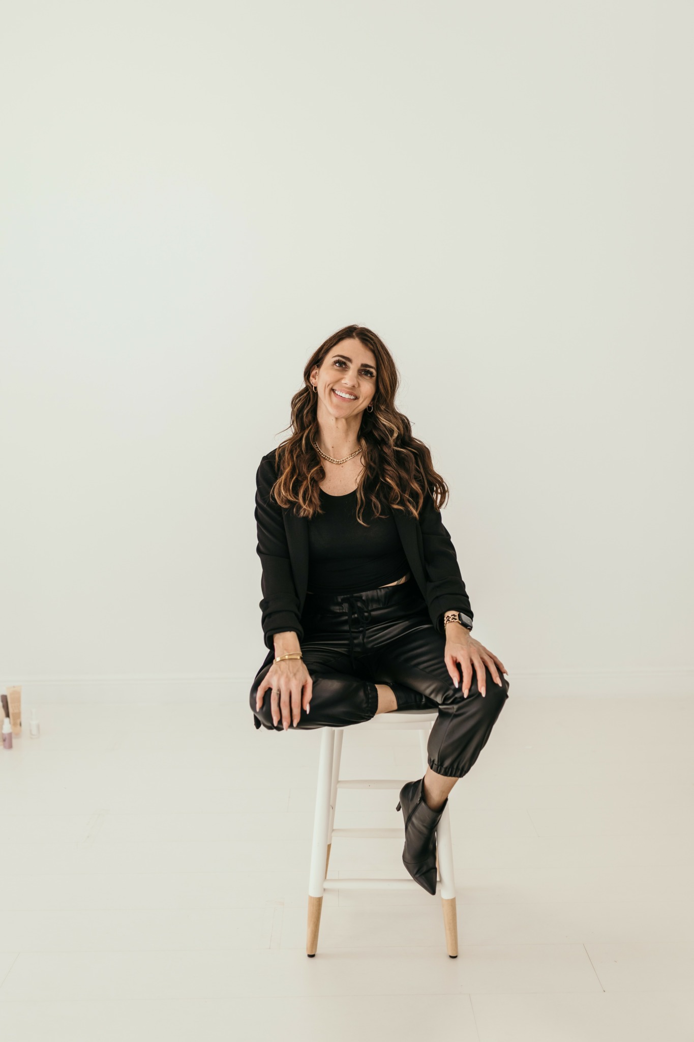 Woman with long wavy hair sitting on a white stool against a plain light background, smiling.