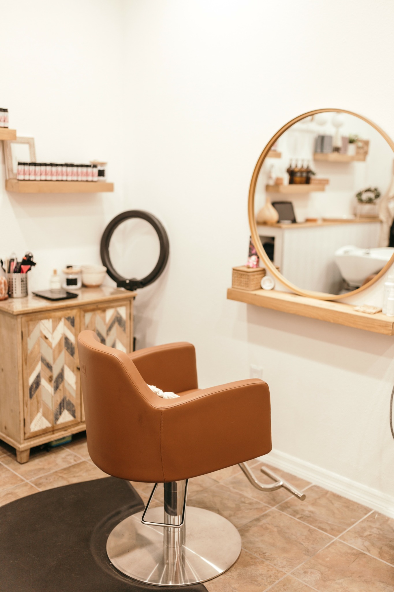 Salon interior with a brown chair, wooden cabinet, round mirror, and wall shelves with beauty products.