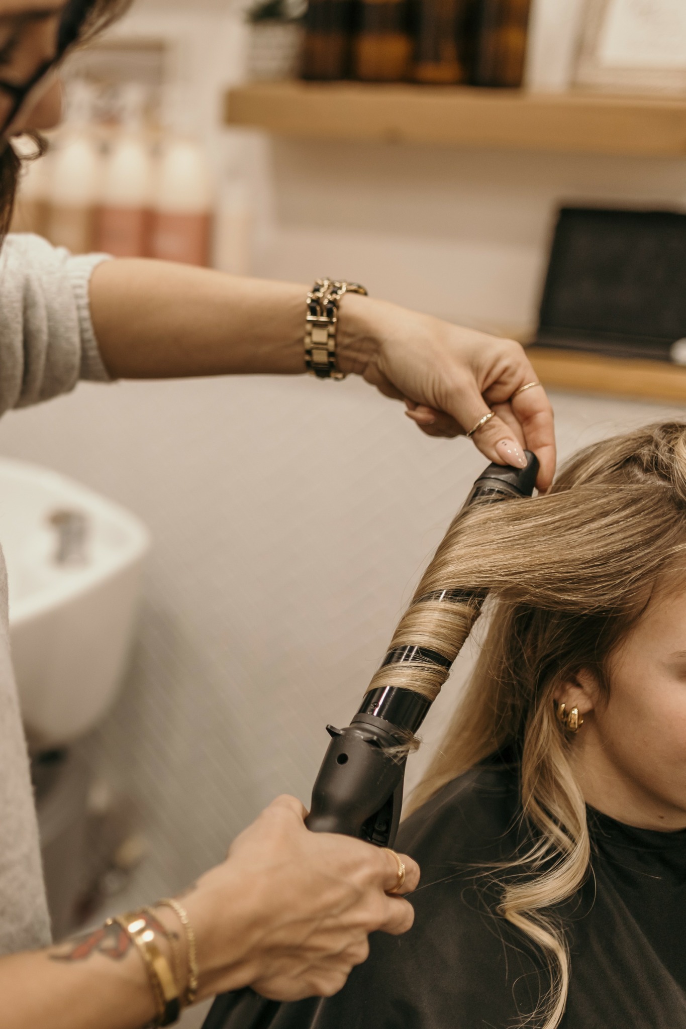 Person styling a woman's hair with a curling iron in a salon setting.
