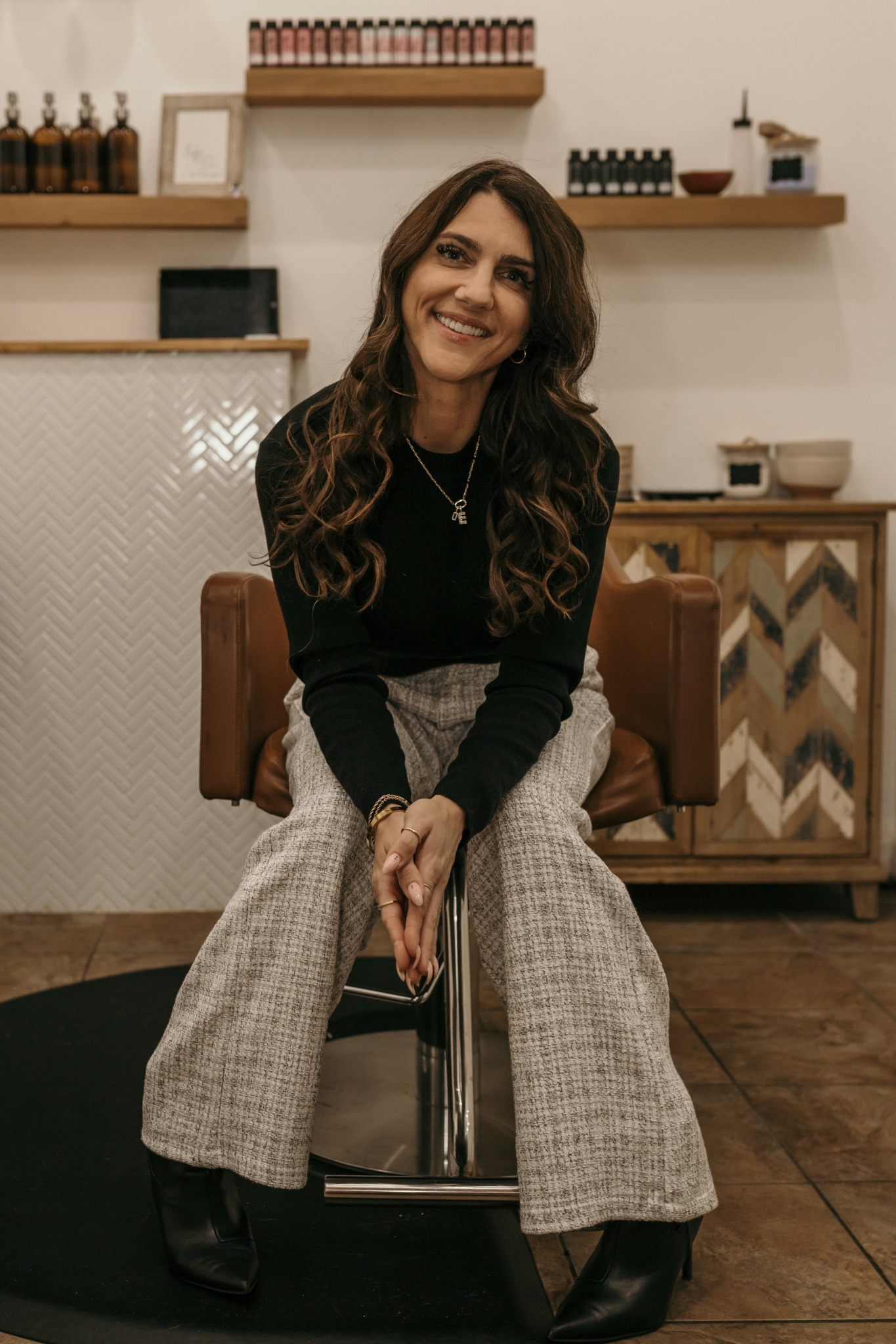 Woman with long wavy hair sitting on a chair, smiling, in a room with shelves and wooden furniture.
