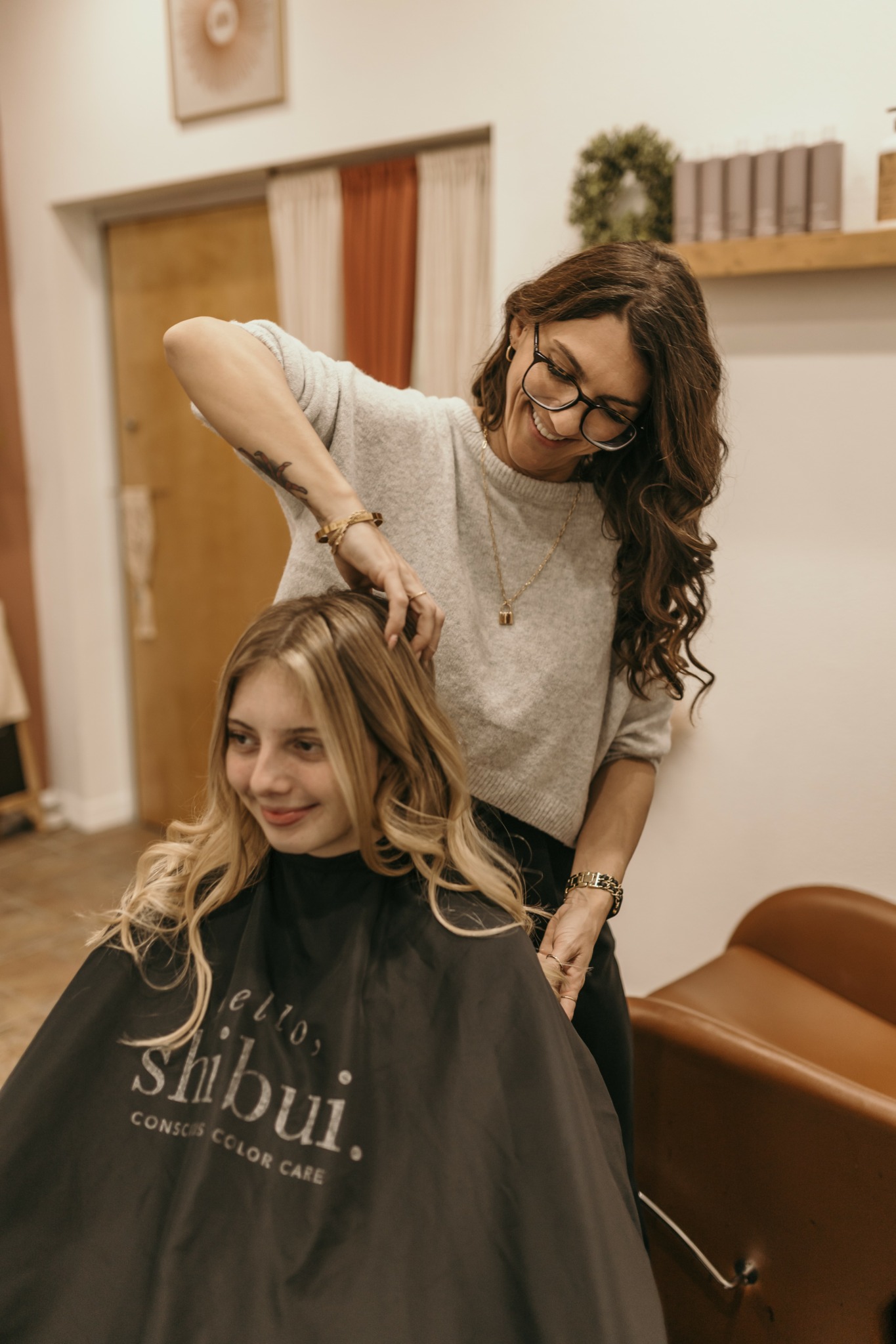 Woman with glasses and long hair cutting a young woman's hair in a salon.