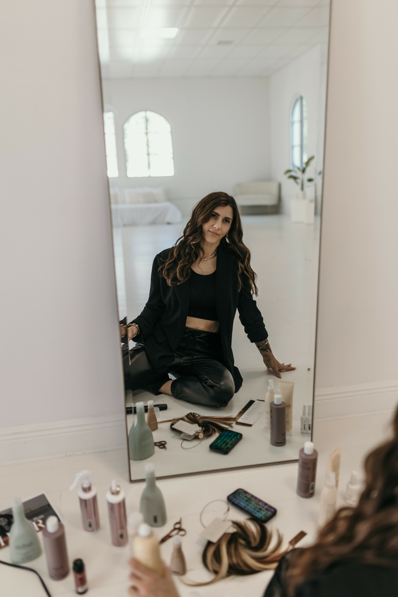 Woman with long hair in black outfit sitting on floor, looking at mirror, surrounded by makeup products.