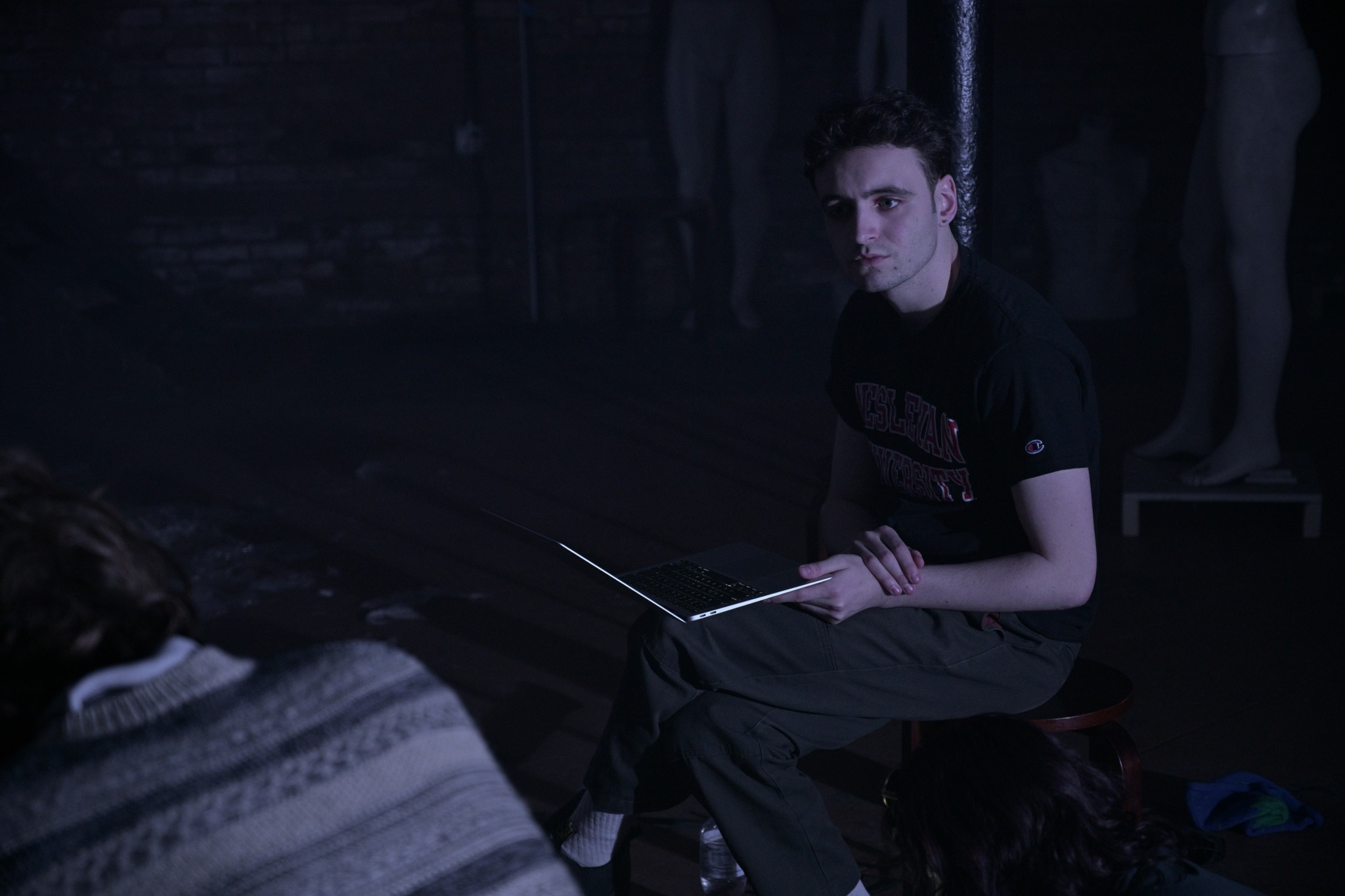 Young man sitting on a stool holding a stick, in a dark room with mannequins in the background.