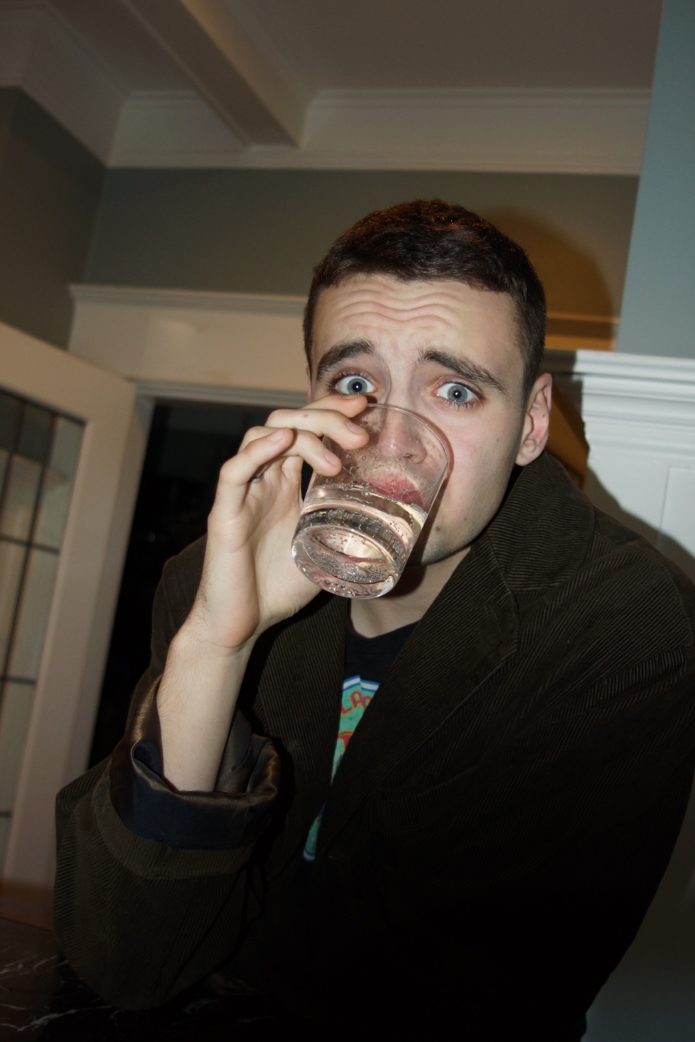 Man with short dark hair holding a glass of water, looking surprised or concerned, indoors.