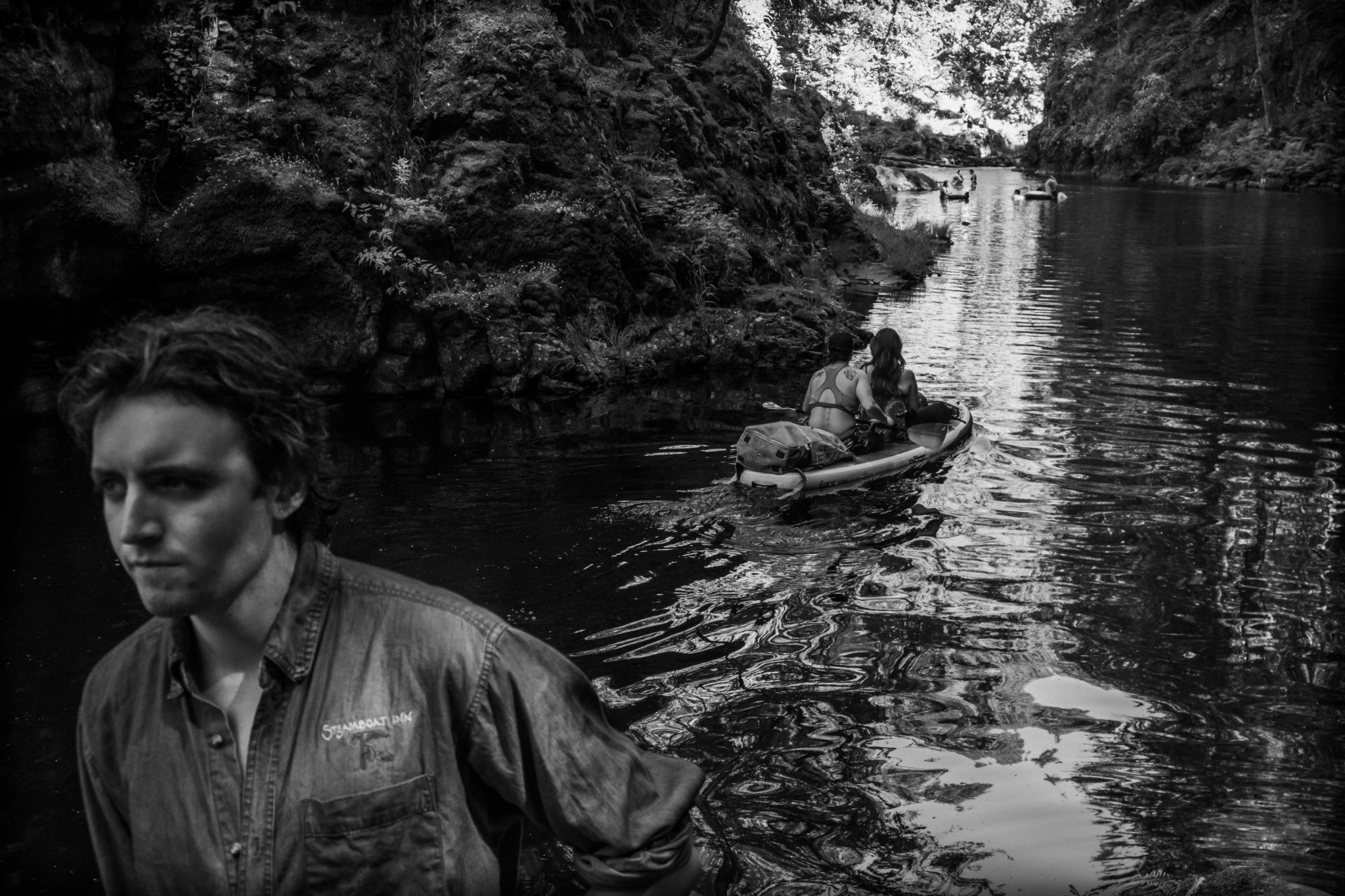 Person in a jacket standing near a river with a person kayaking in the background, surrounded by rocky cliffs.