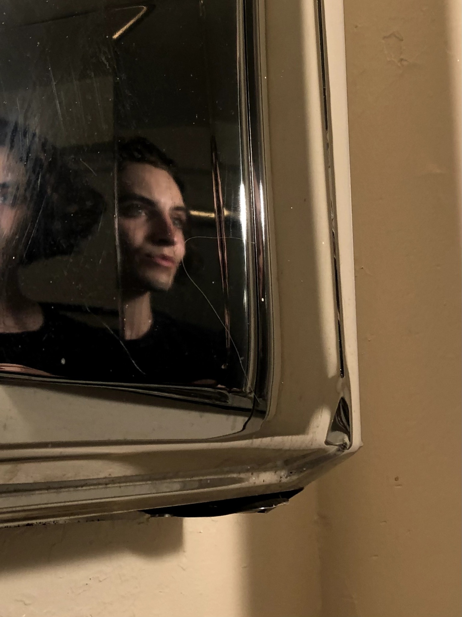 Person's face reflected in a mirror or glass surface, with dark background and warm lighting.