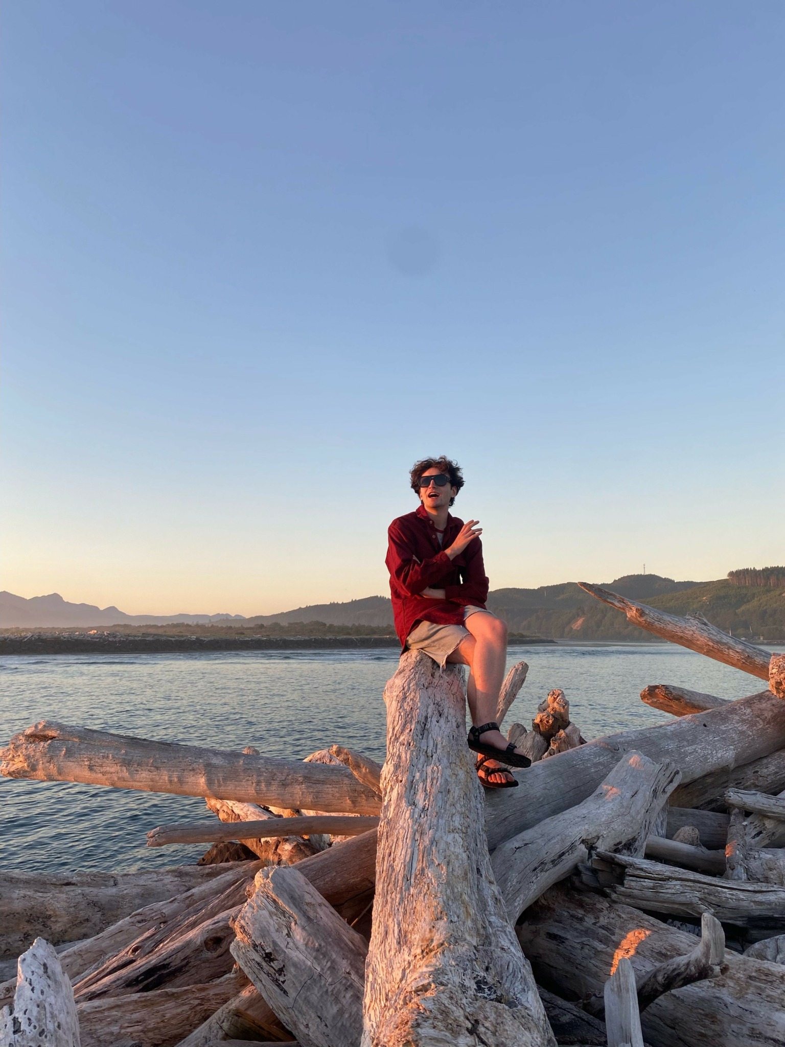 Person sitting on a large log near a body of water during sunset, with a clear sky and distant horizon.
