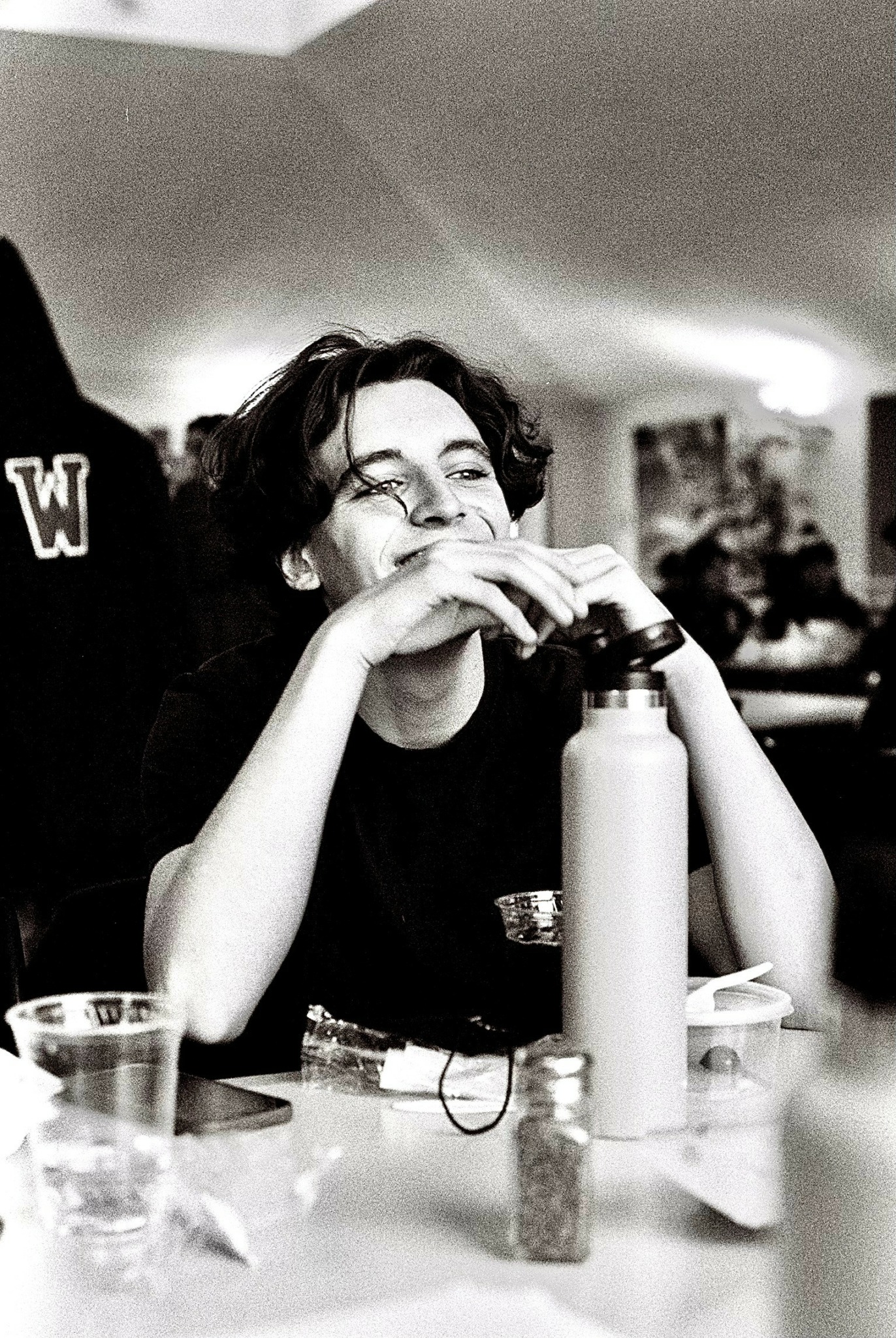Person with curly hair smiling, sitting at a table with a water bottle and glasses, in a room with other people.