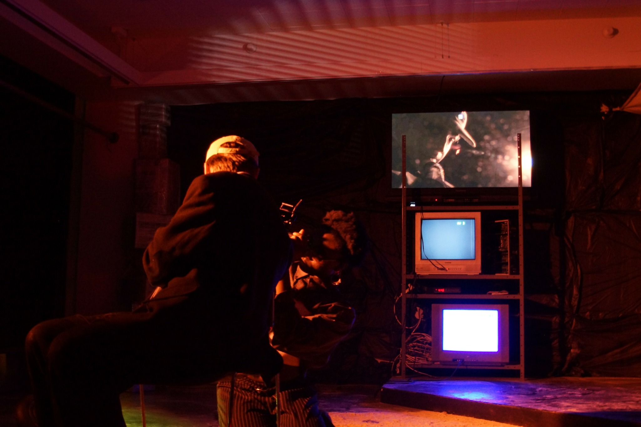 Person performing on stage with screens displaying a musician, surrounded by equipment, in a dimly lit setting.