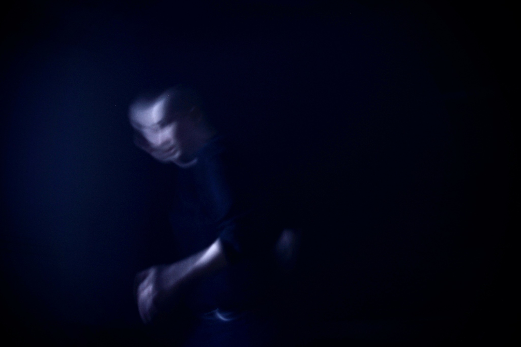 Person with glasses in a dark environment, face illuminated slightly, looking downward.
