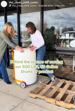 Two women move a large white container on a yellow hand truck outside a building with glass windows.