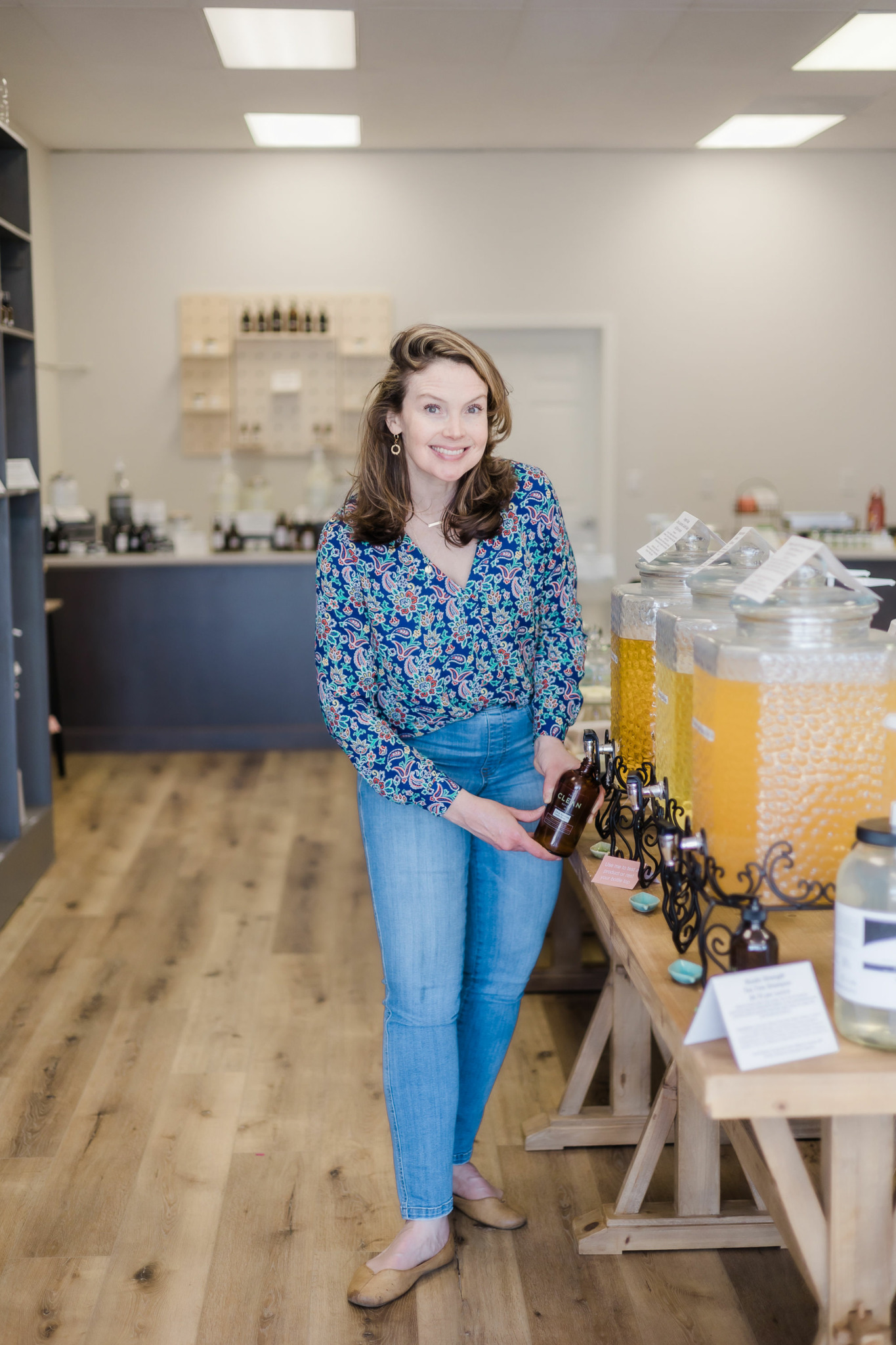 Young woman with wavy hair in a floral blouse and jeans stands in a store with wooden tables and containers.