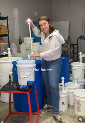 Woman in white hoodie and jeans standing next to cleaning supplies and buckets in a room.