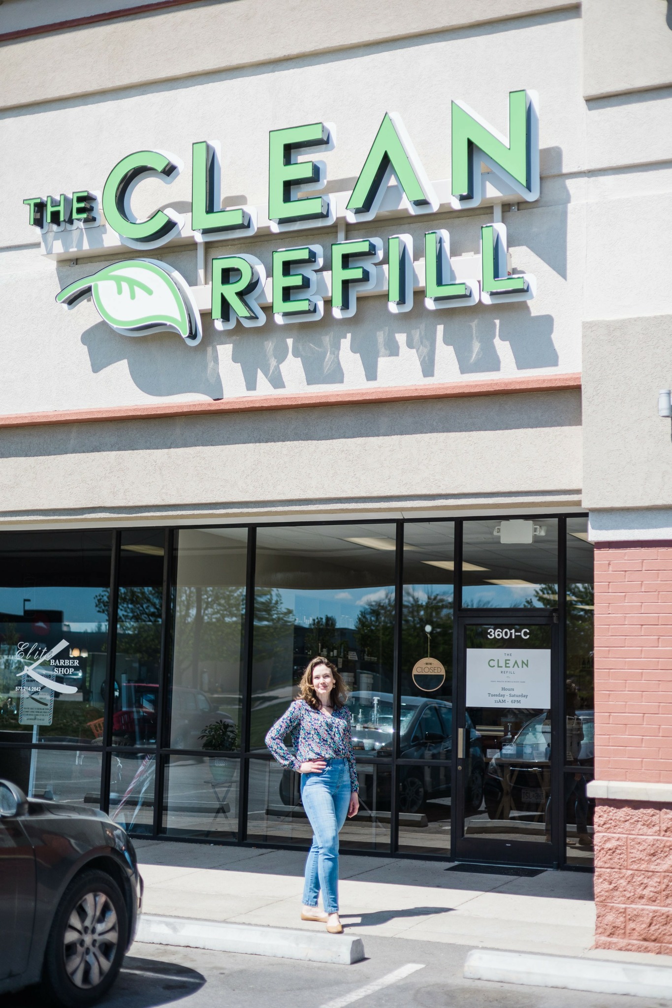 Person standing outside refill shop with glass windows and sign reading 'The Clean Refill'.