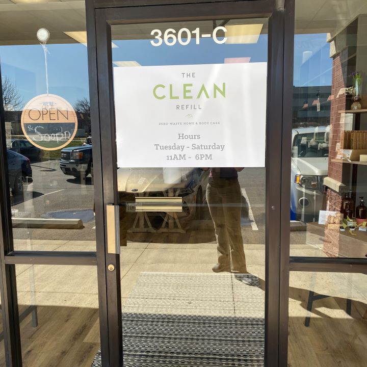 Glass door with store hours and sign, reflection of person and parking lot outside, store interior visible.